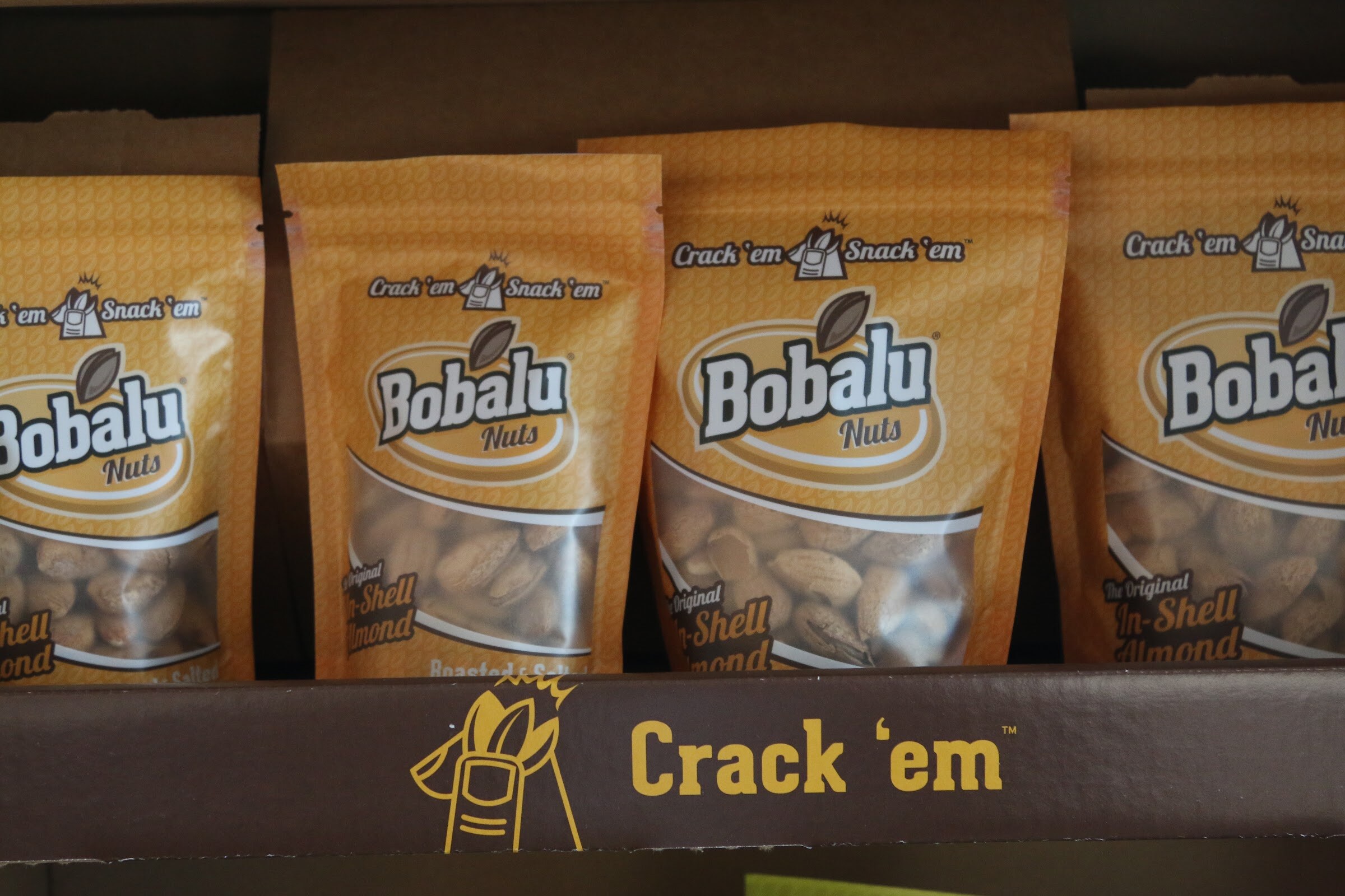 Gold Bobalu Nuts pouches of in-shell almonds standing in a brown tray printed with Crack 'em.