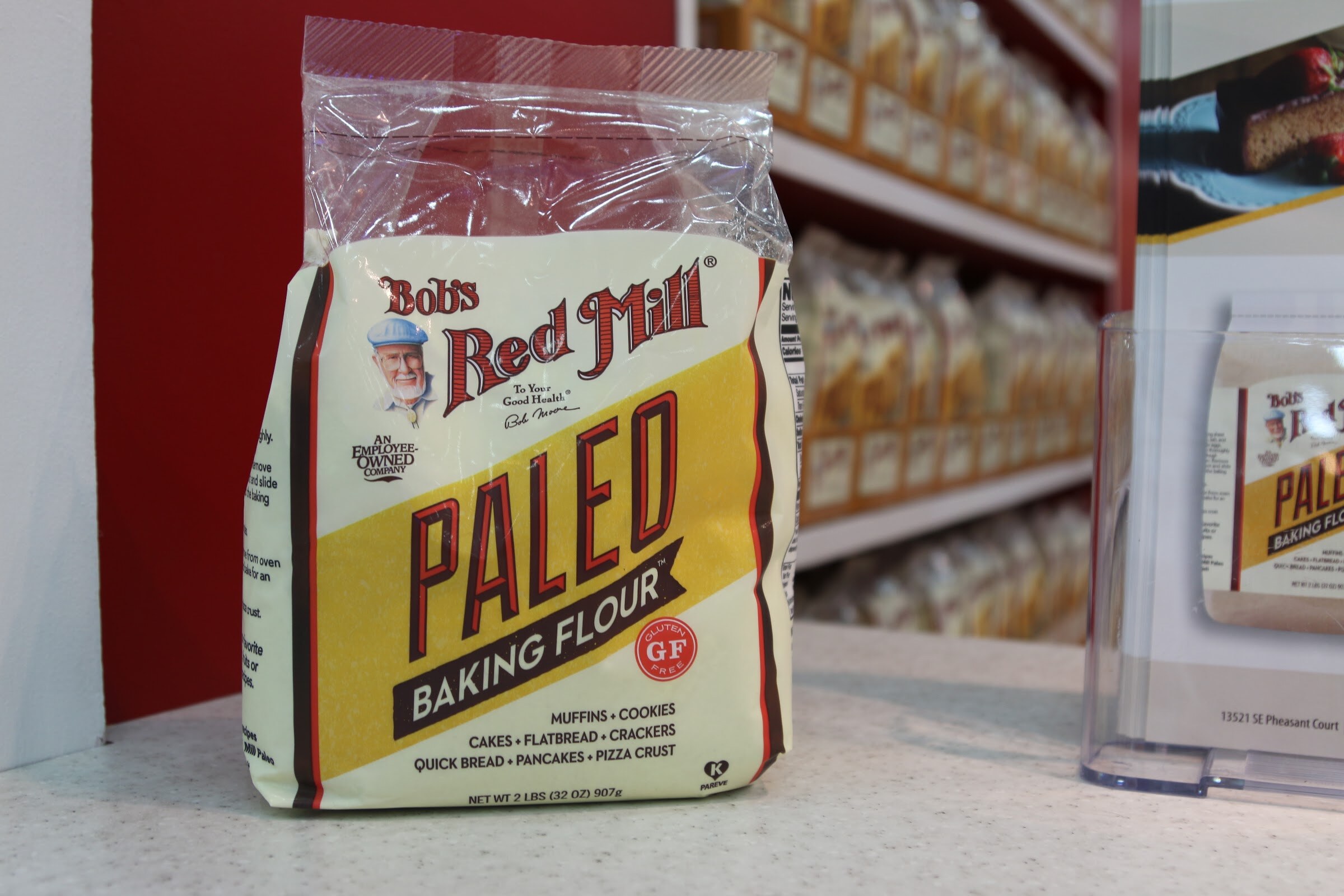 A single bag of Bob's Red Mill Paleo Baking Flour in front of shelves of Bob's Red Mill products and a red wall.