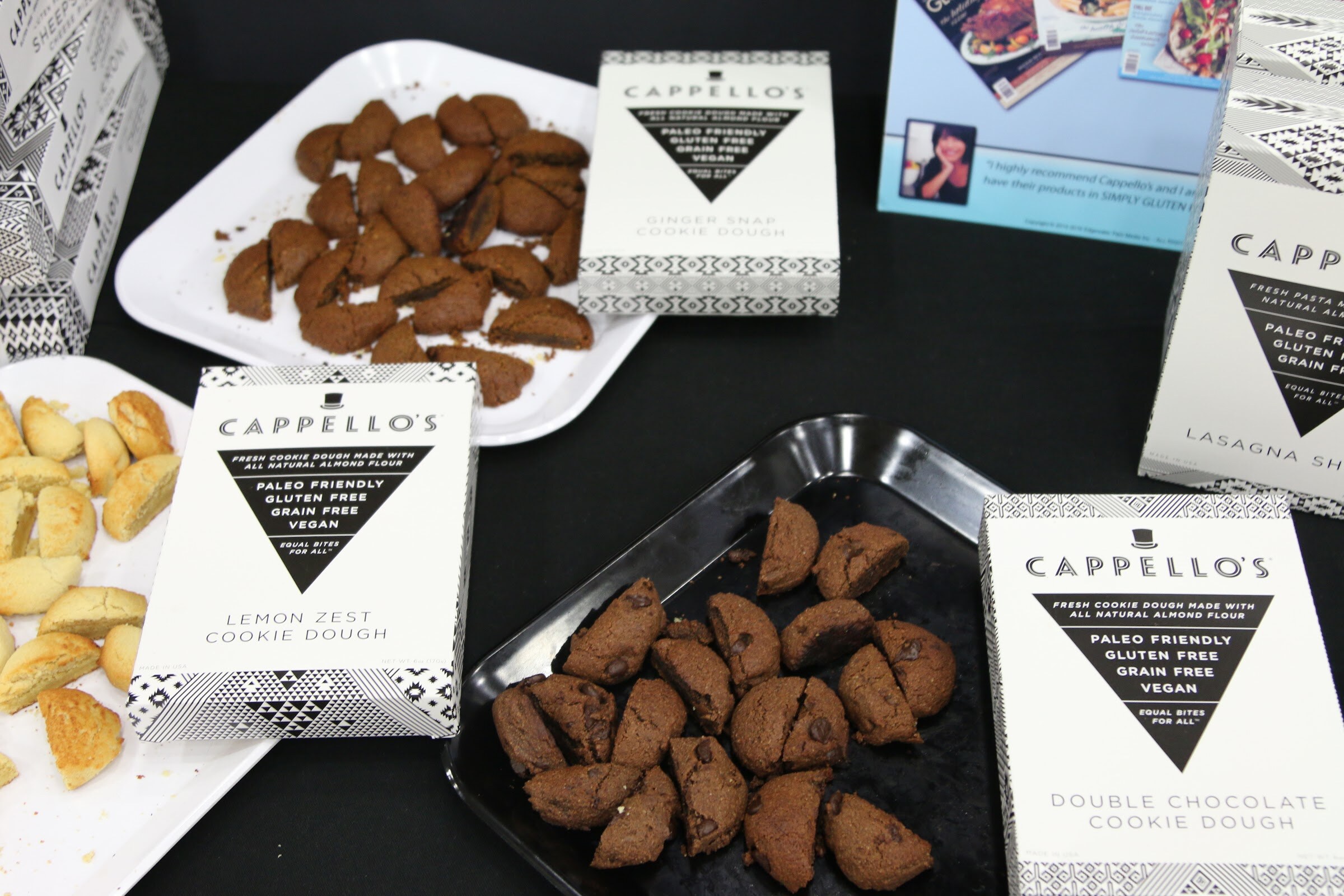 White Cappello's cookie dough boxes in lemon zest, ginger snap and double chocolate on a black table next to trays of sample cookies.