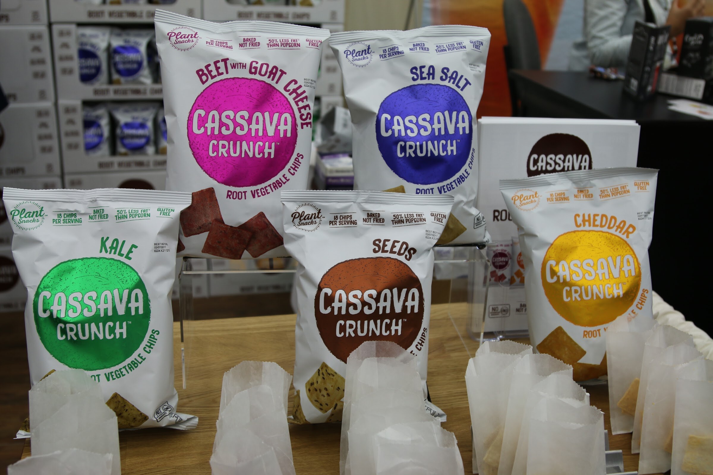 Five colorful Cassava Crunch root vegetable chip bags on an acrylic stand with small paper sample bags in front.