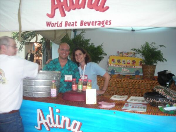 A man in a teal patterned shirt and a woman in a light blue shirt smile behind the Adina World Beat Beverages booth counter, with juice bottles in front and a man in white facing them.