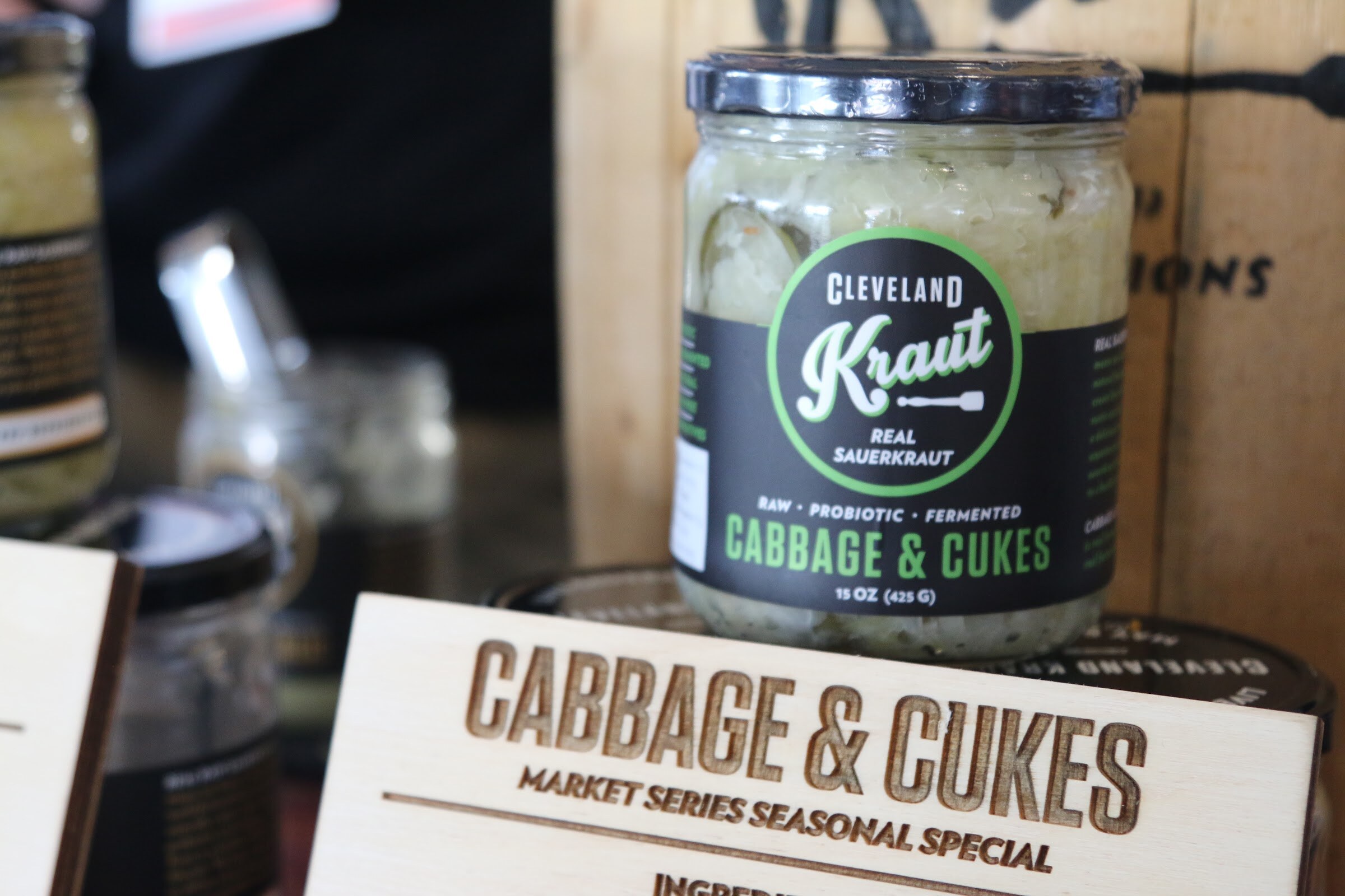 A Cleveland Kraut real sauerkraut jar with a black and green label above an engraved wood sign reading Cabbage and Cukes.