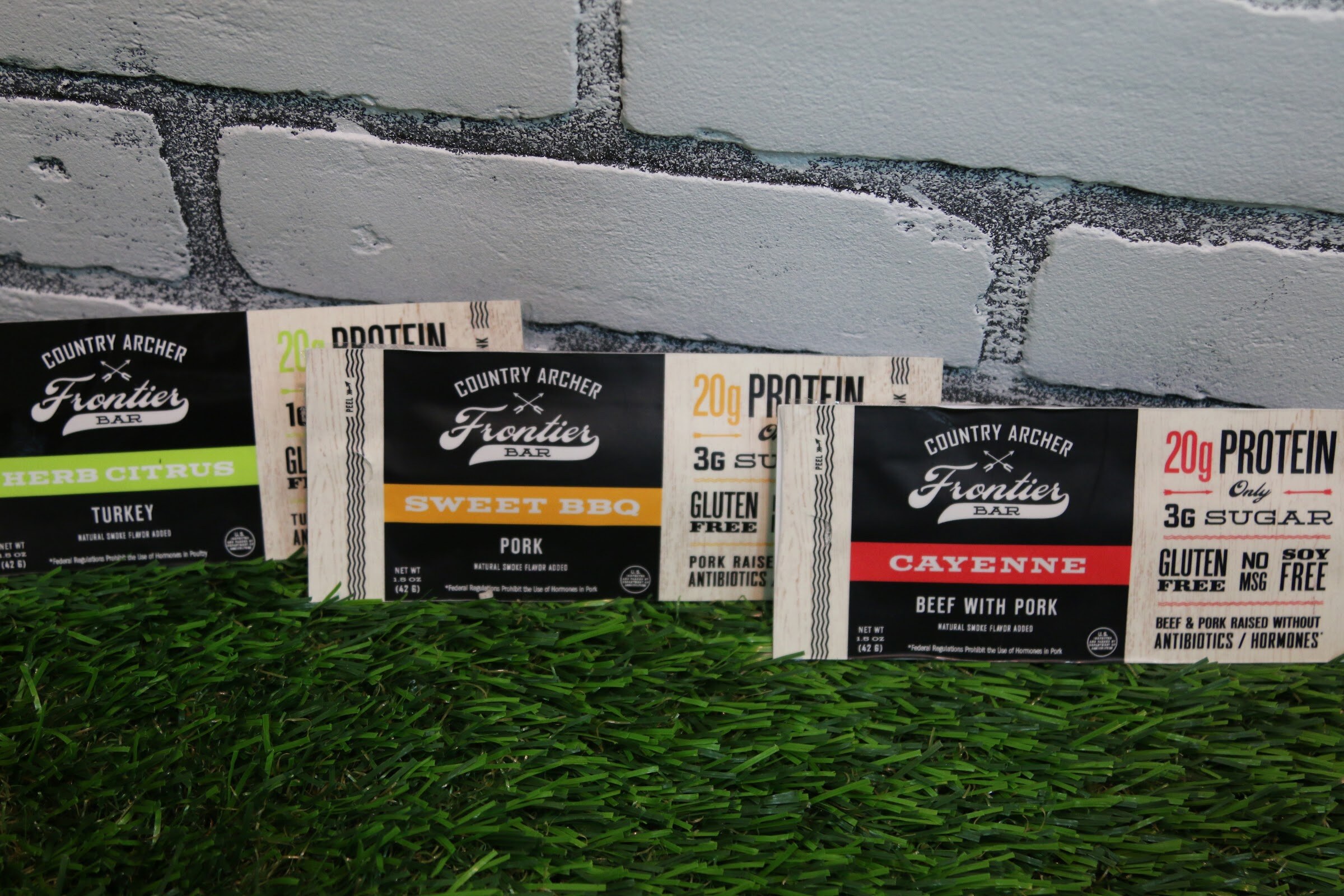 Three Country Archer Frontier Bar packs leaning against a painted white brick wall on a strip of green artificial grass.