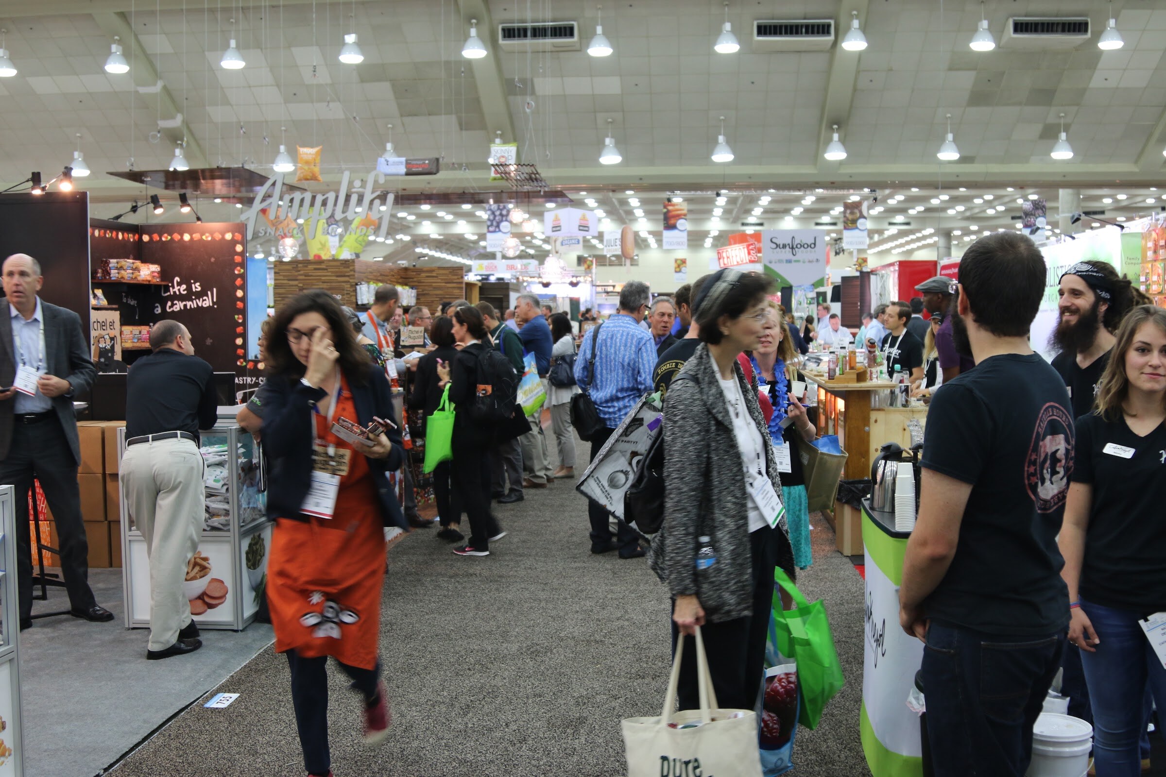 Busy convention aisle with a woman in an orange apron walking past attendees holding green tote bags and exhibitors at booths.