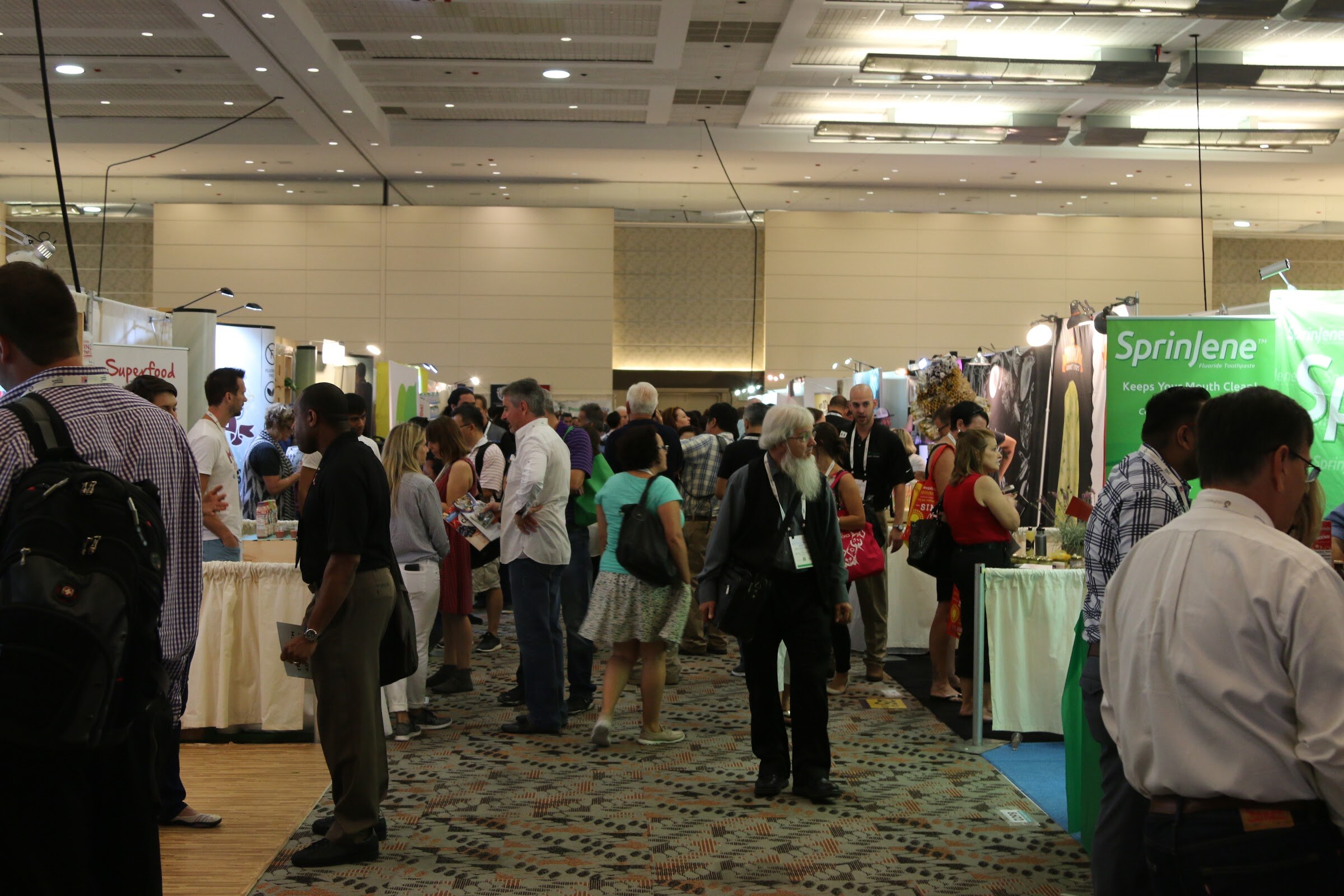 A busy aisle in a convention hall ceiling lit with recessed lights, with men and women in lanyards walking between booths including a green Sprinjene banner.