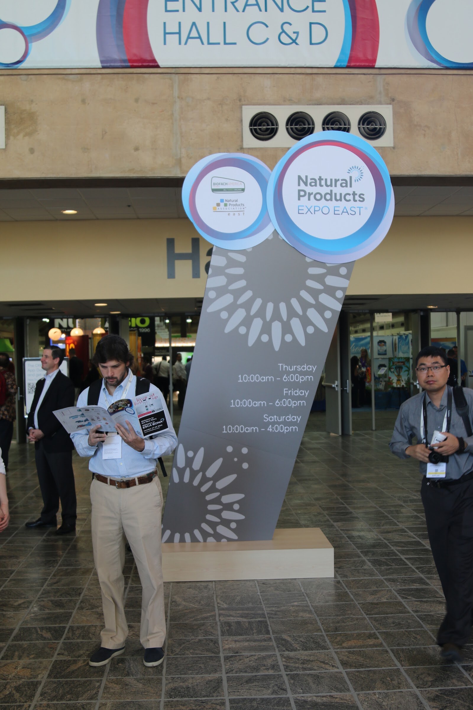 A man reads a program beside a tall gray Natural Products Expo East sign below a Hall C and D entrance banner, with another attendee at right.