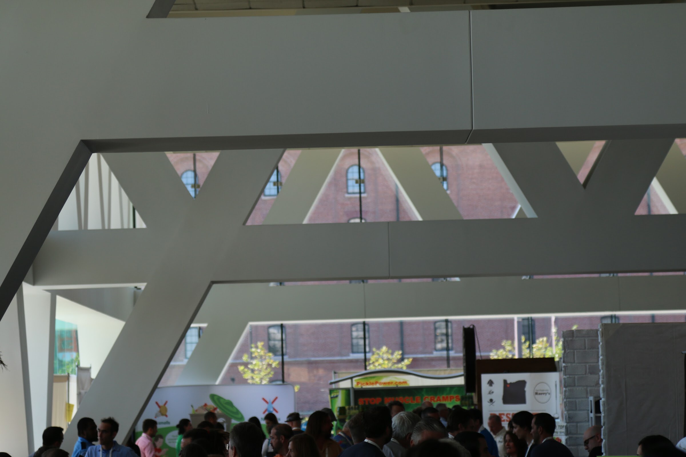 View up at a white angular steel truss structure with a crowd of attendees below and a brick building in the background.