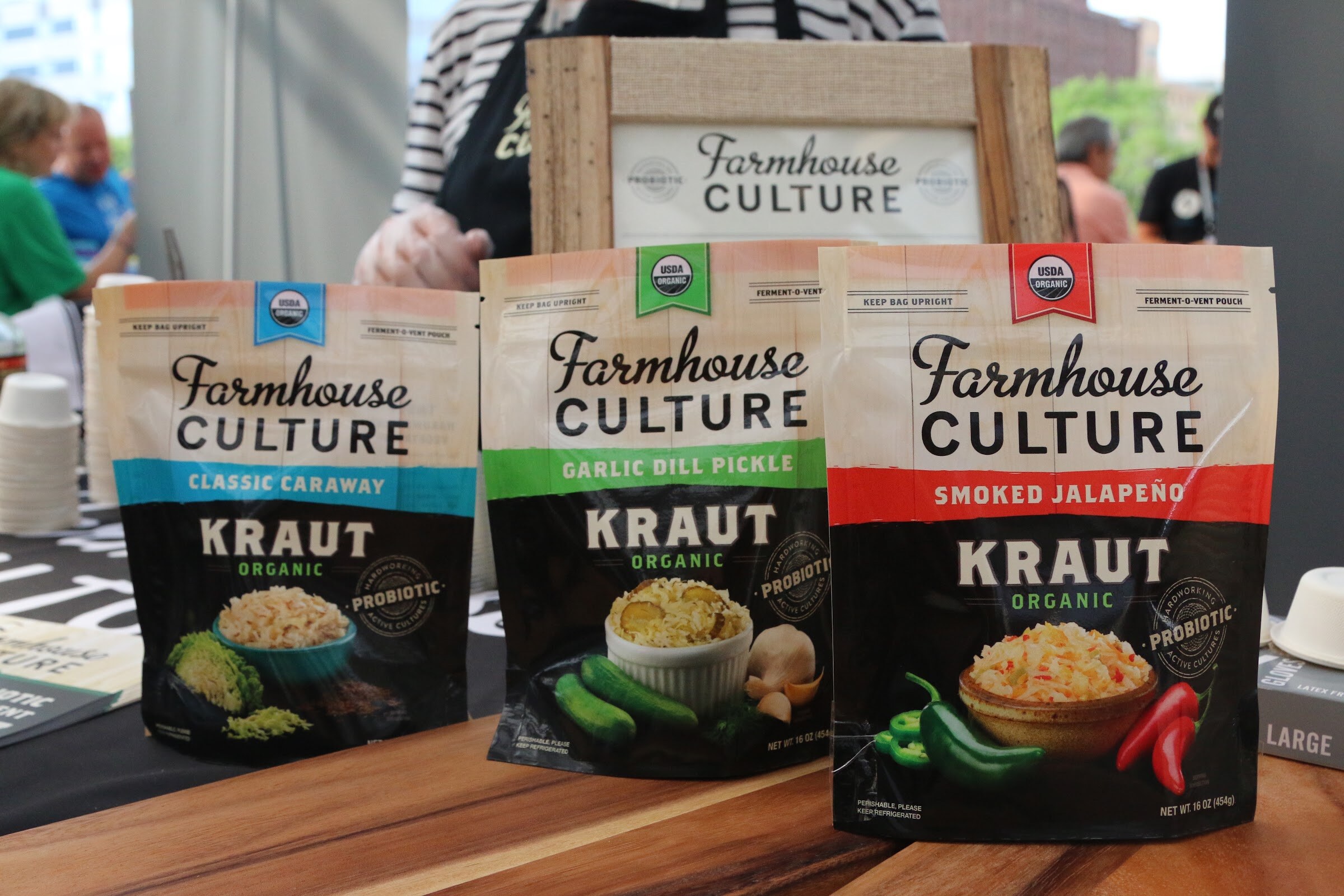 Three Farmhouse Culture kraut pouches in classic caraway, garlic dill pickle and smoked jalapeno on a wooden board.