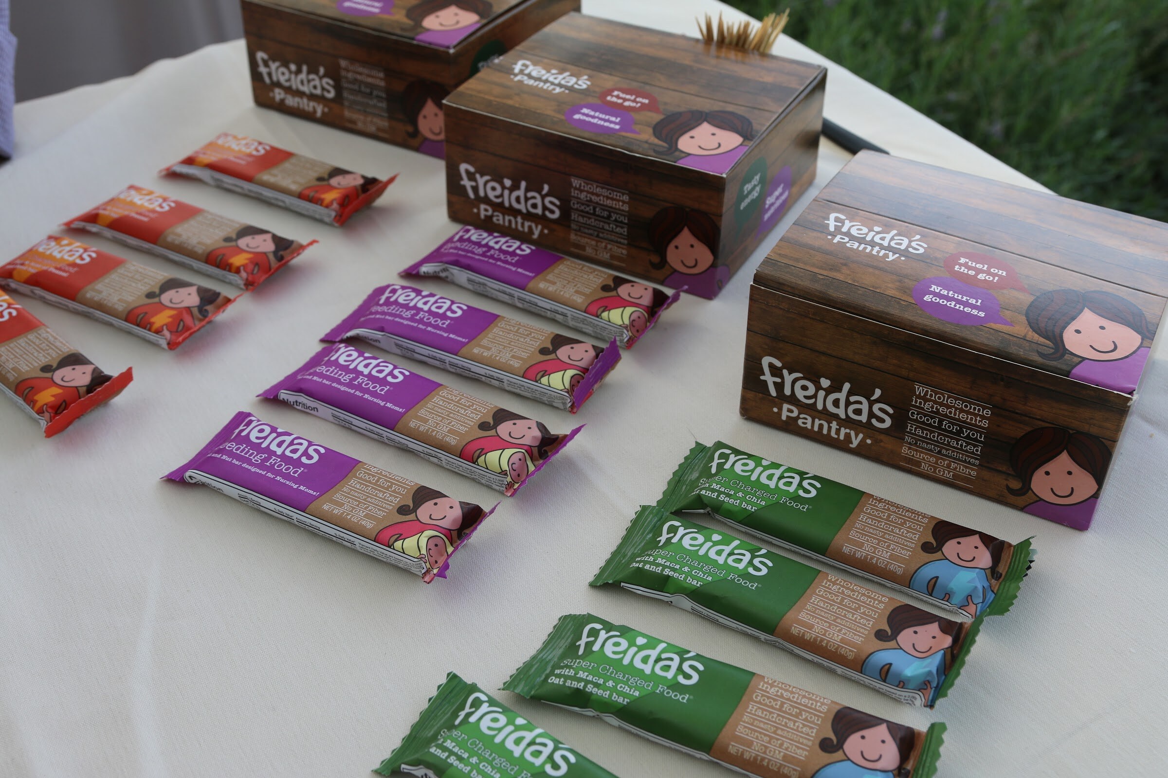 Freida's Pantry bars fanned out on a white tablecloth next to wooden Freida's Pantry boxes.