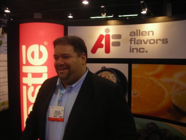 A smiling man in a dark blazer and badge stands in the Allen Flavors Inc. booth, with the company logo, orange slices imagery and a red Nestle banner behind him.