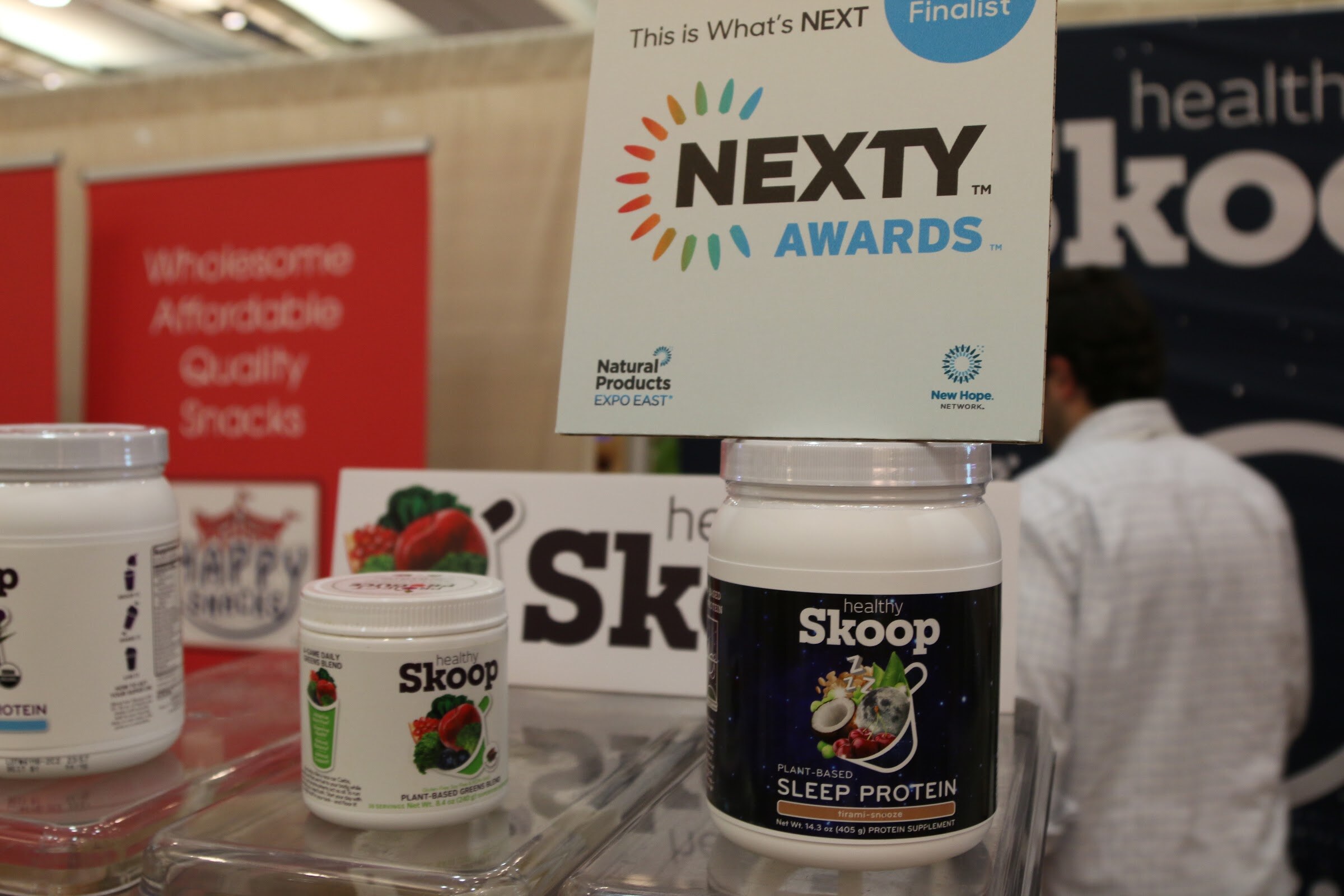 A white NEXTY Awards finalist card above Healthy Skoop sleep protein tubs on a clear stand, with a man in a white shirt behind.