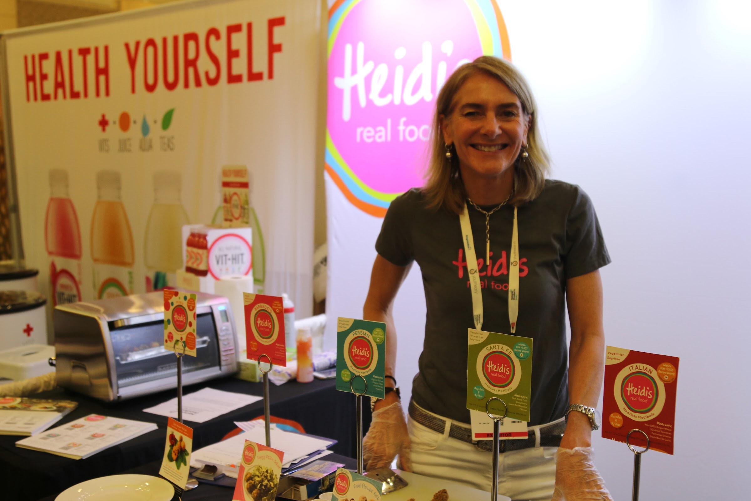 A smiling woman in a gray shirt with a lanyard stands behind a table with food samples under a Health Yourself banner and a pink Heidi's sign.