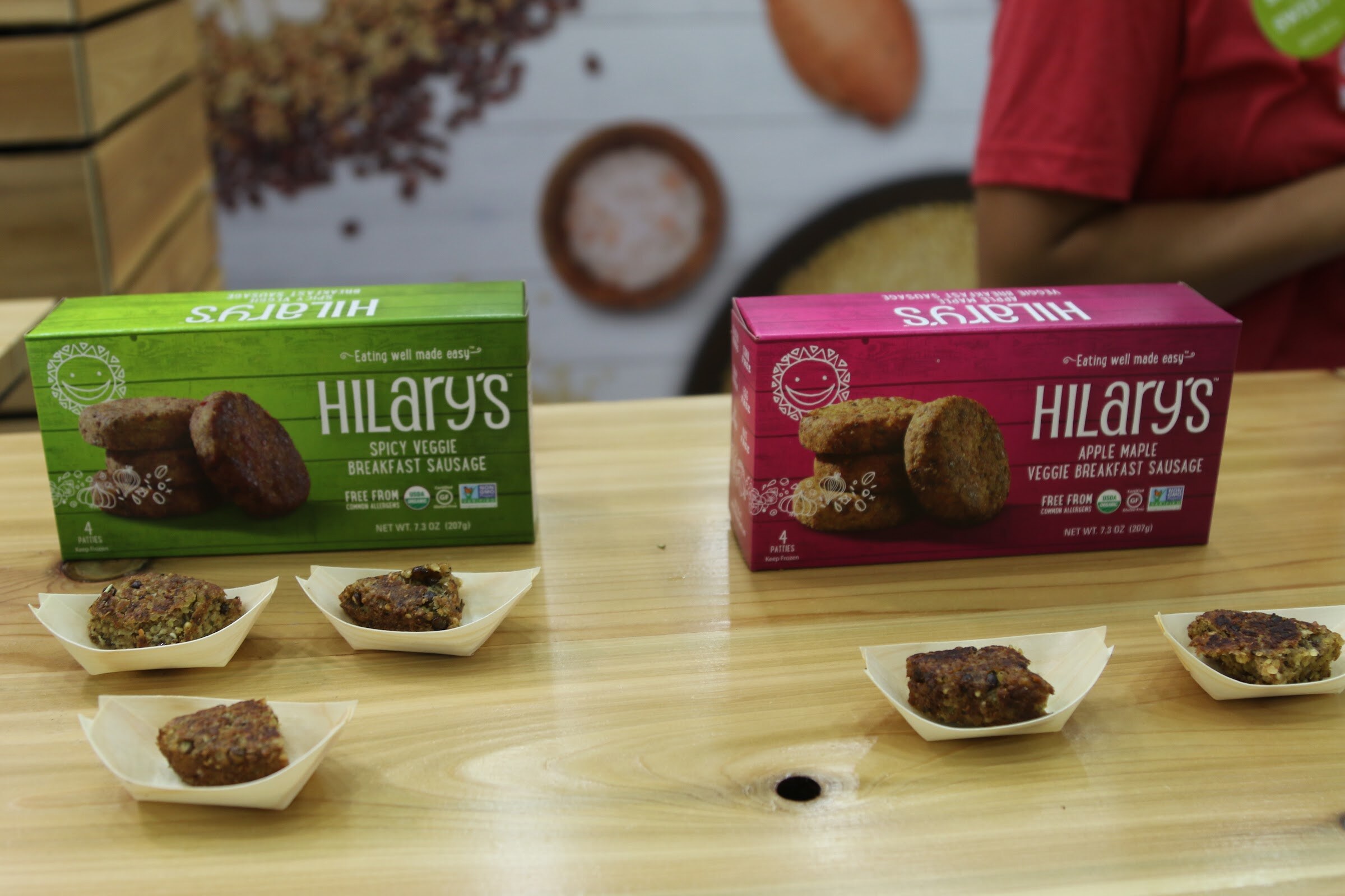 Green and pink Hilary's veggie breakfast sausage boxes on a wood counter with four small boats of sample bites in front.
