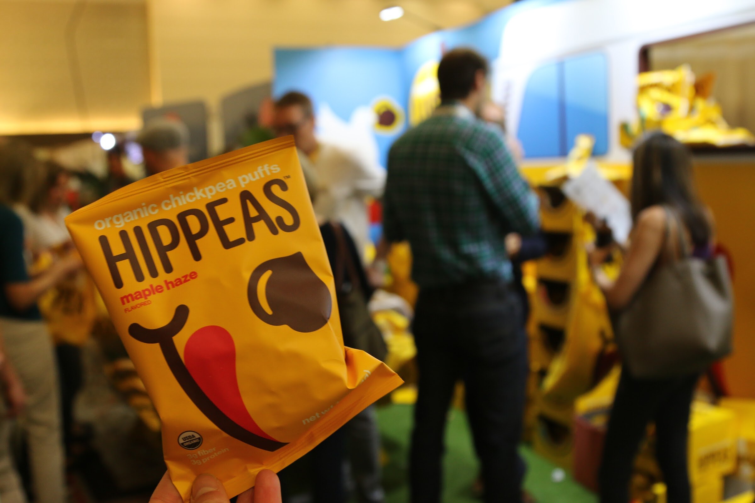 A bright yellow Hippeas chickpea puffs bag in the foreground with blurred visitors and yellow displays behind it.