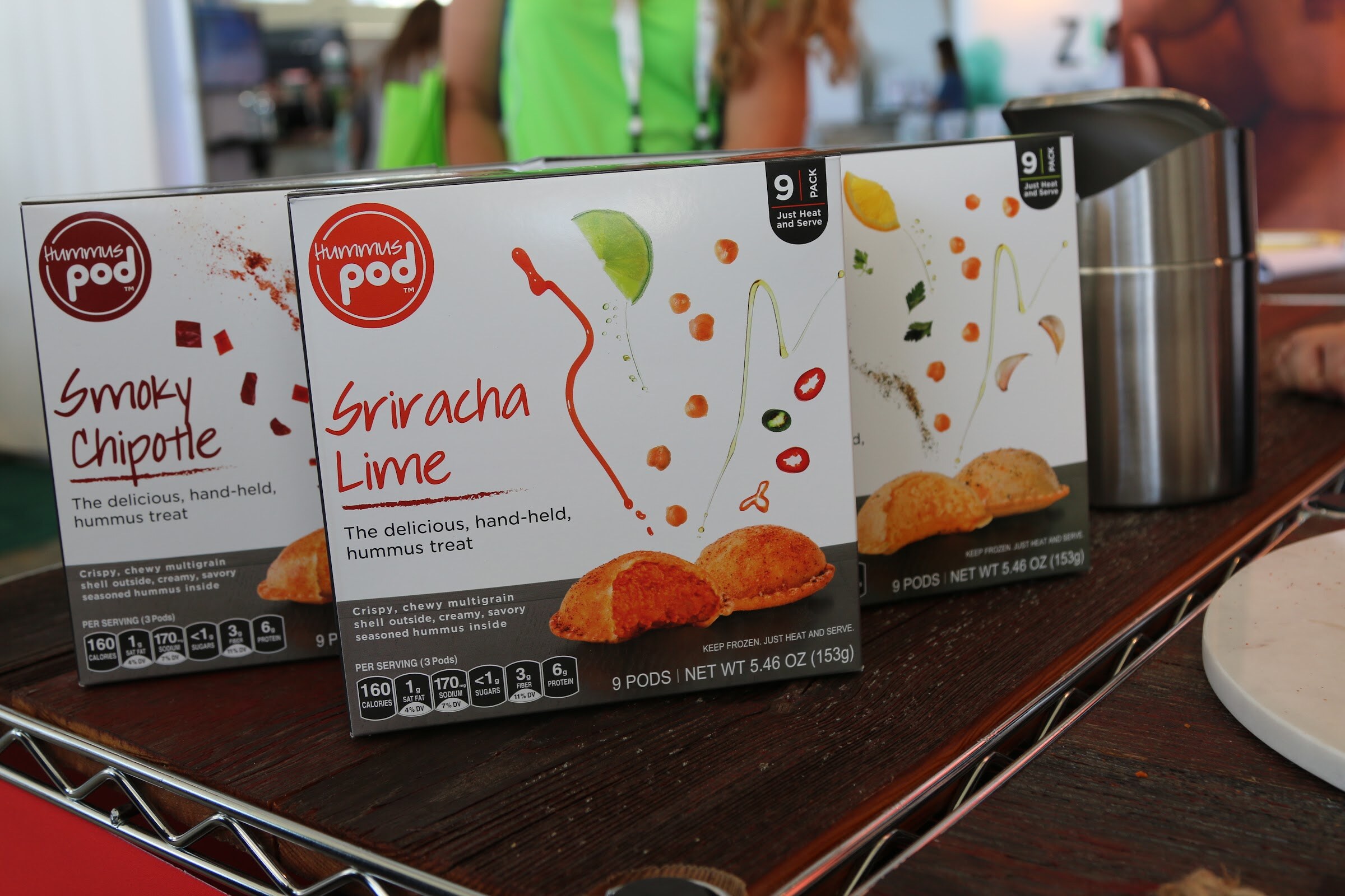 Three white Hummus Pod boxes with images of baked hummus pastries on a dark wood shelf, with people in green shirts blurred behind.