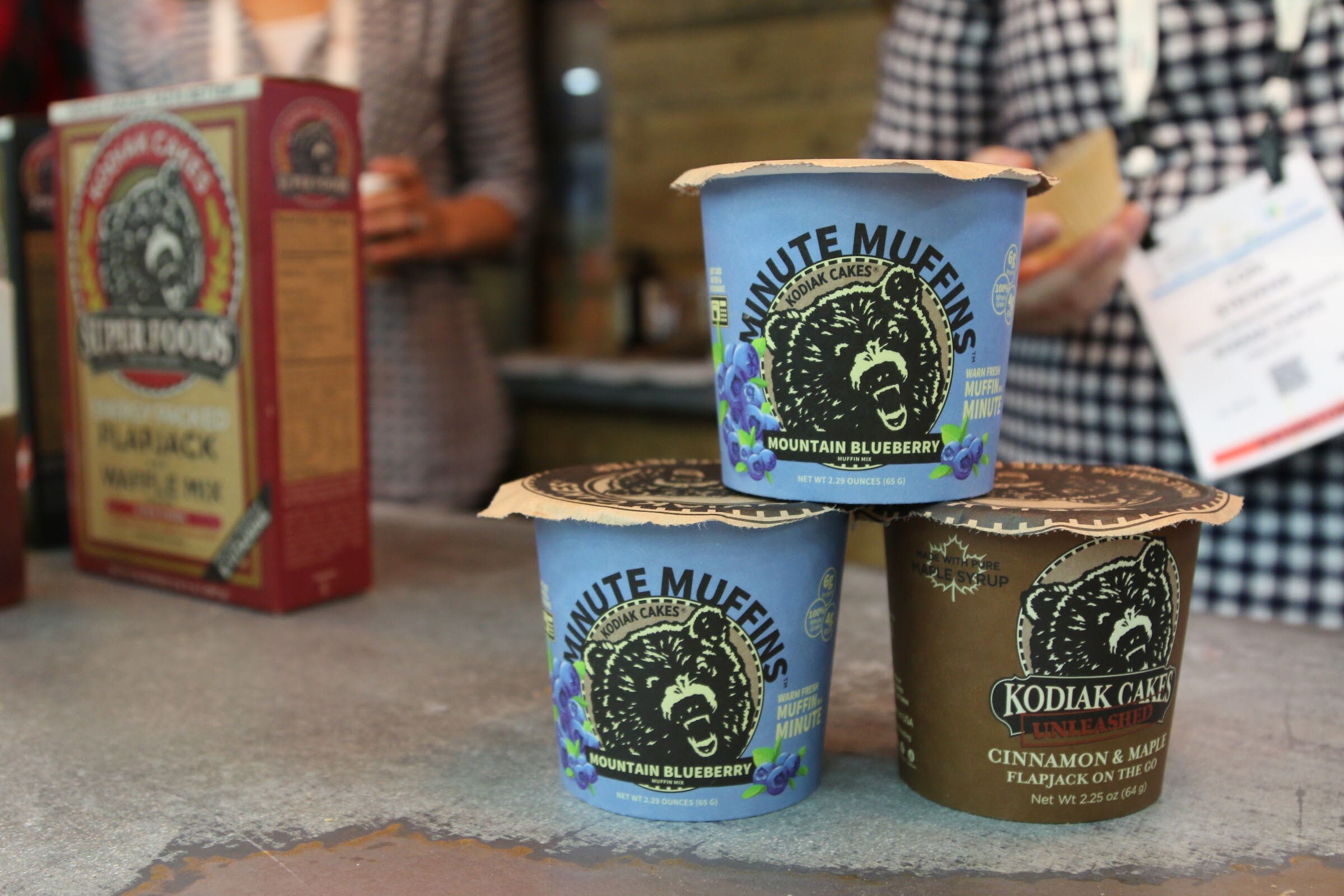 Three Kodiak Cakes cups with bear logos stacked on a gray table in front of a Kodiak Super Foods box, with an attendee in checks behind.