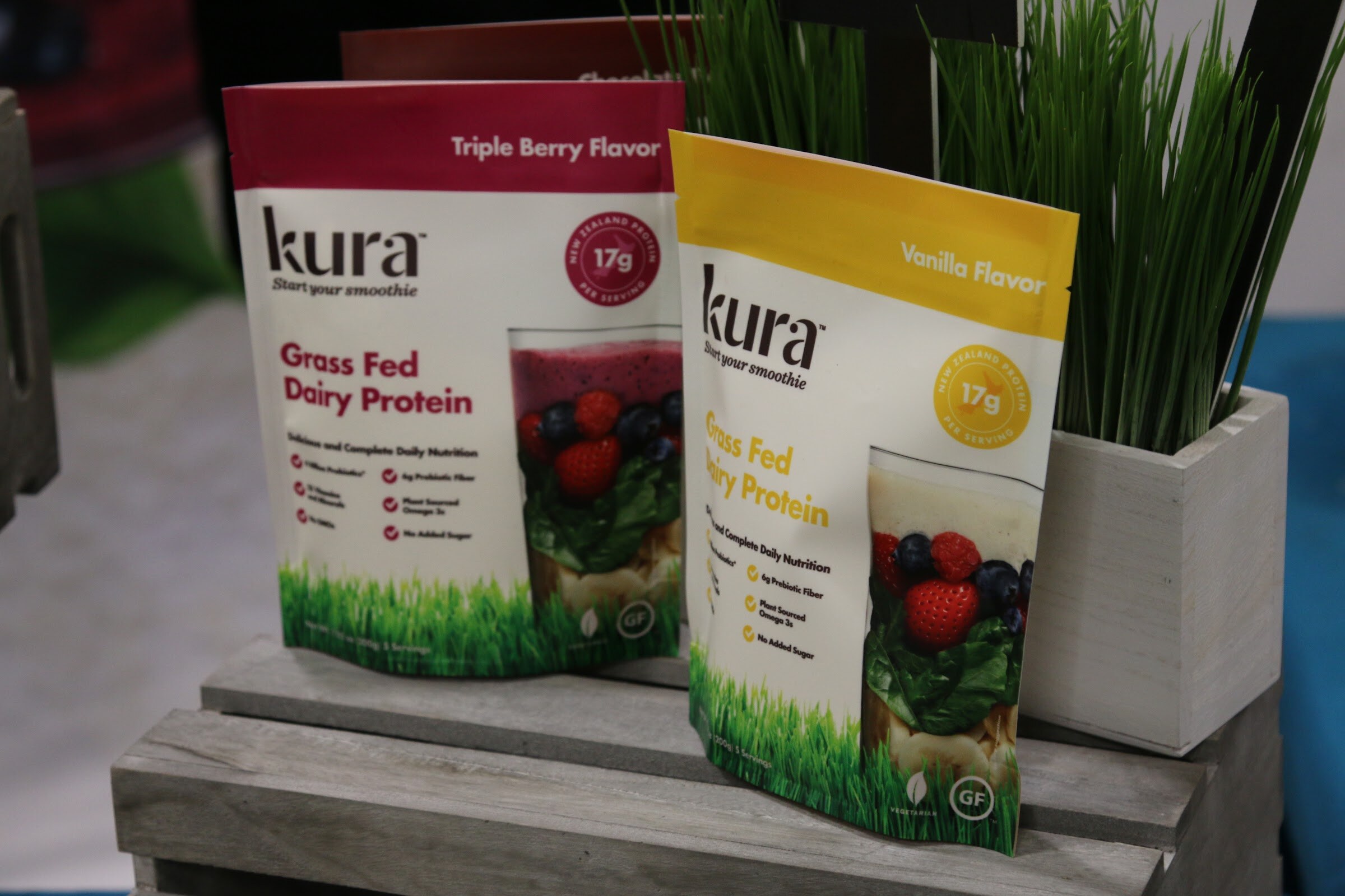 Two Kura smoothie pouches with fruit photos on a gray wooden crate with a small wheatgrass planter.