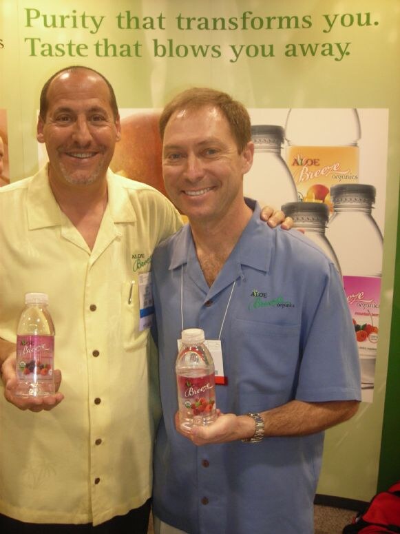 Two smiling men, one in a yellow shirt and one in a blue shirt with Aloe Breeze logos, each hold a bottle of Aloe Breeze in front of a display reading Purity that transforms you.