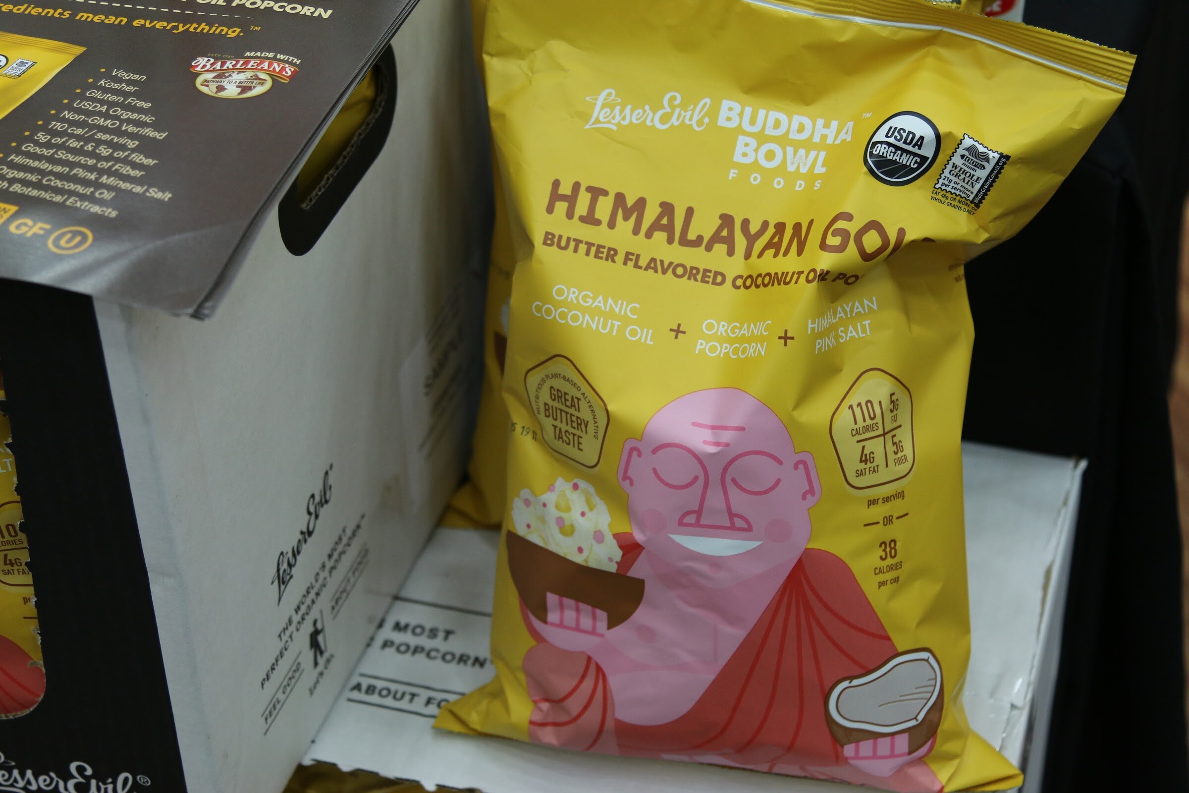 A yellow Lesser Evil Buddha Bowl Himalayan Gold popcorn bag with a smiling pink buddha illustration next to a white cardboard display.