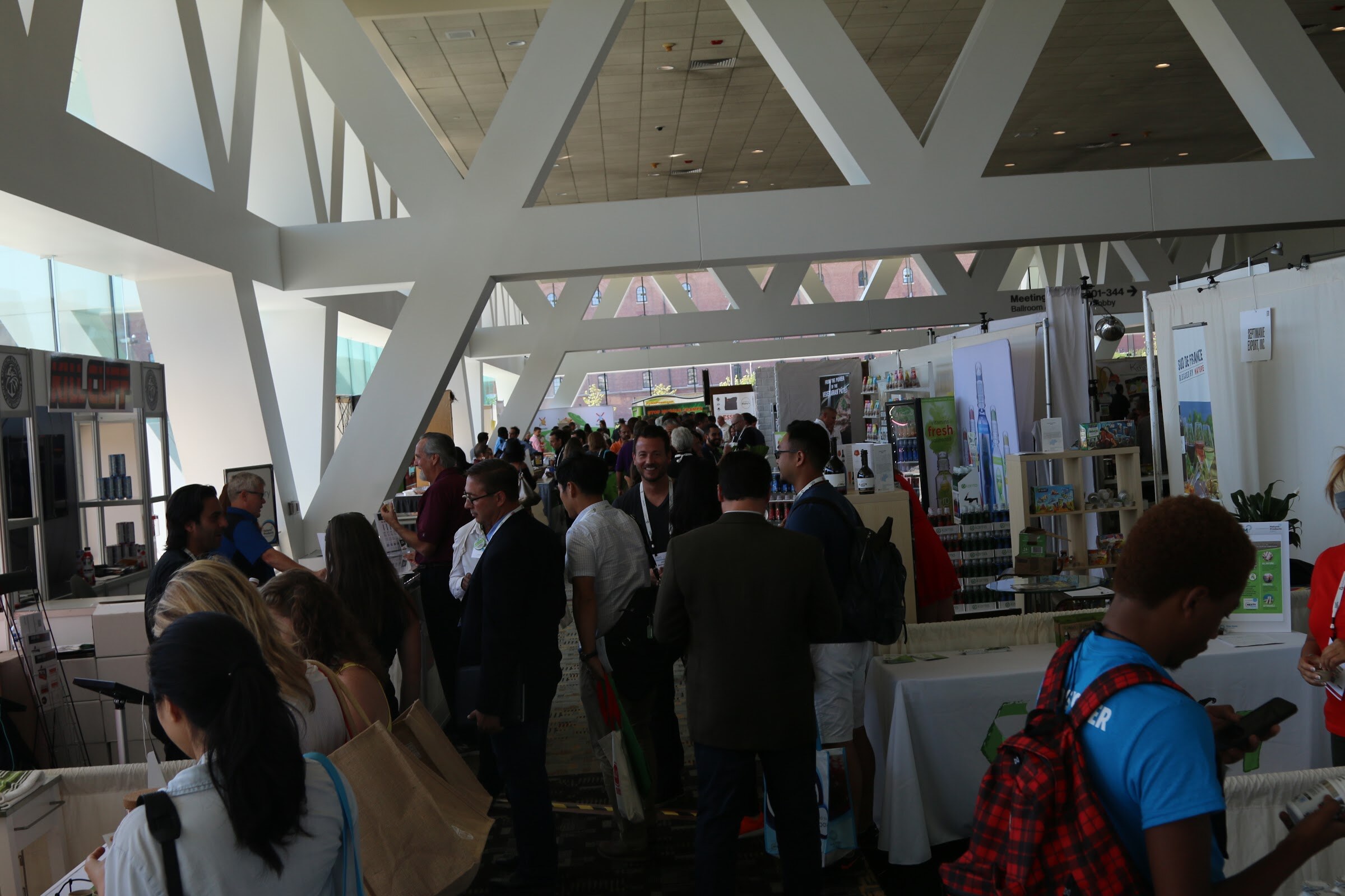 Dense crowd of attendees in a long hall with white geometric ceiling trusses, with one man in a blue shirt and red backpack checking his phone in the foreground.