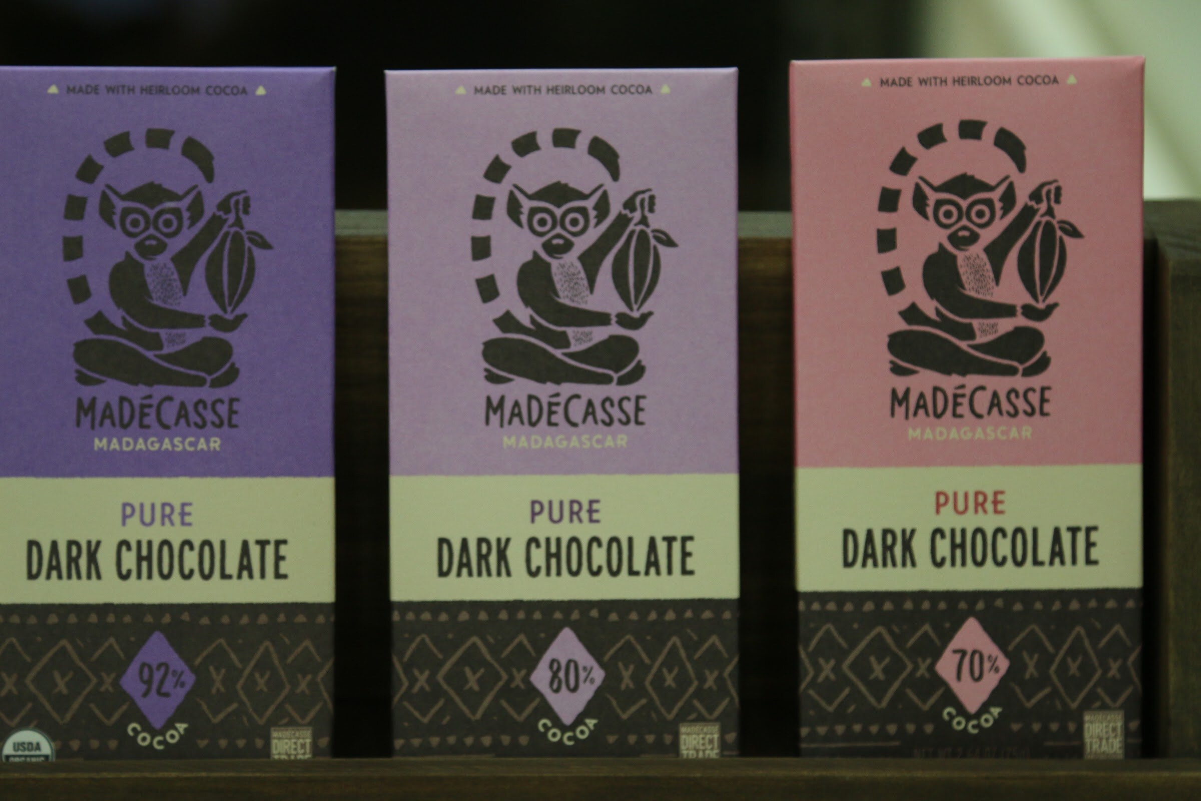 Three Madecasse chocolate bars with lemur illustrations in purple, lavender and pink wrappers on a dark wooden shelf.