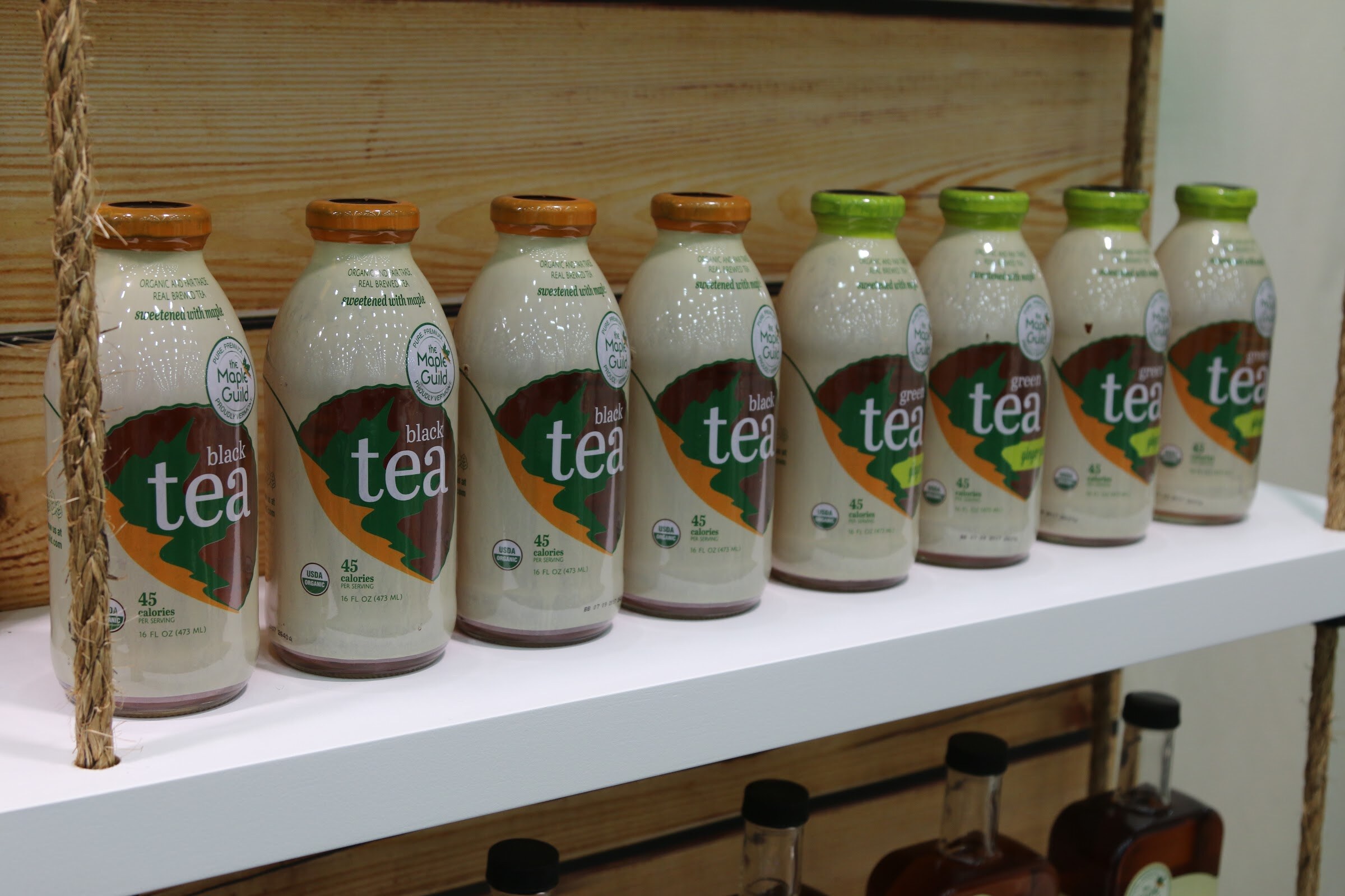 Eight milky-colored bottles labeled tea in orange and green caps on a white shelf, with maple syrup bottles on the shelf below.