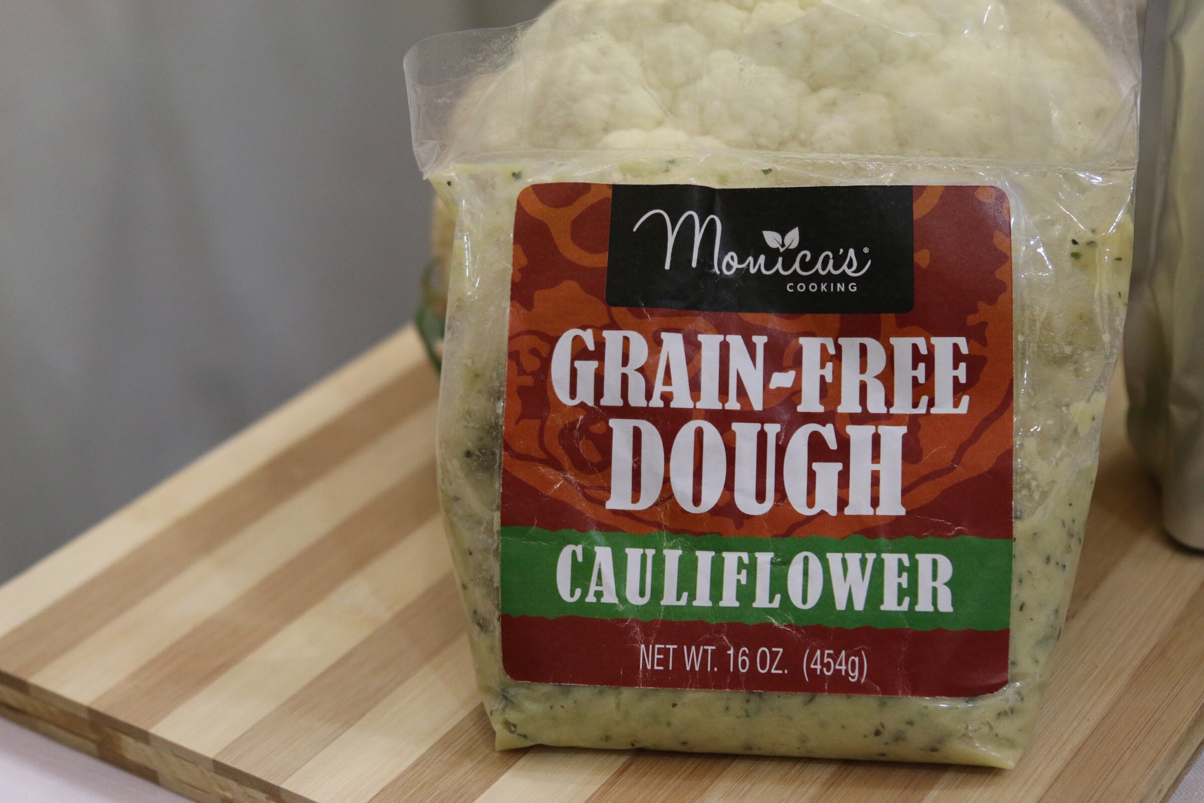 A clear bag of cauliflower dough with a red and green Monica's Cooking label on a striped wood cutting board.