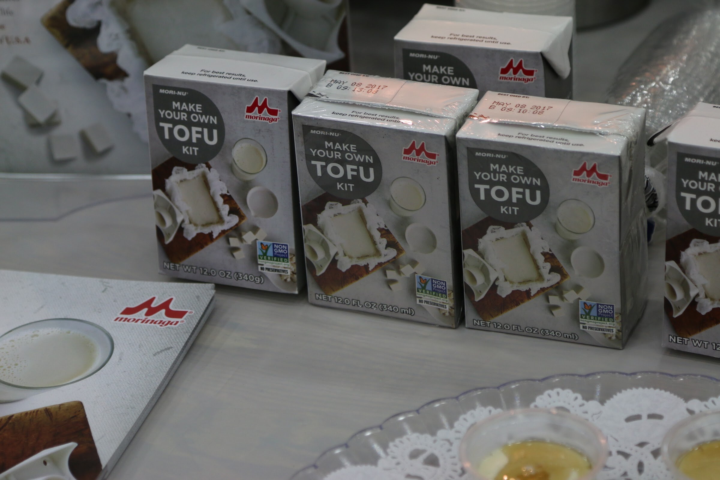 Three gray Morinaga Mori-Nu Make Your Own Tofu Kit boxes on a table beside a booklet and a clear tray holding small samples.