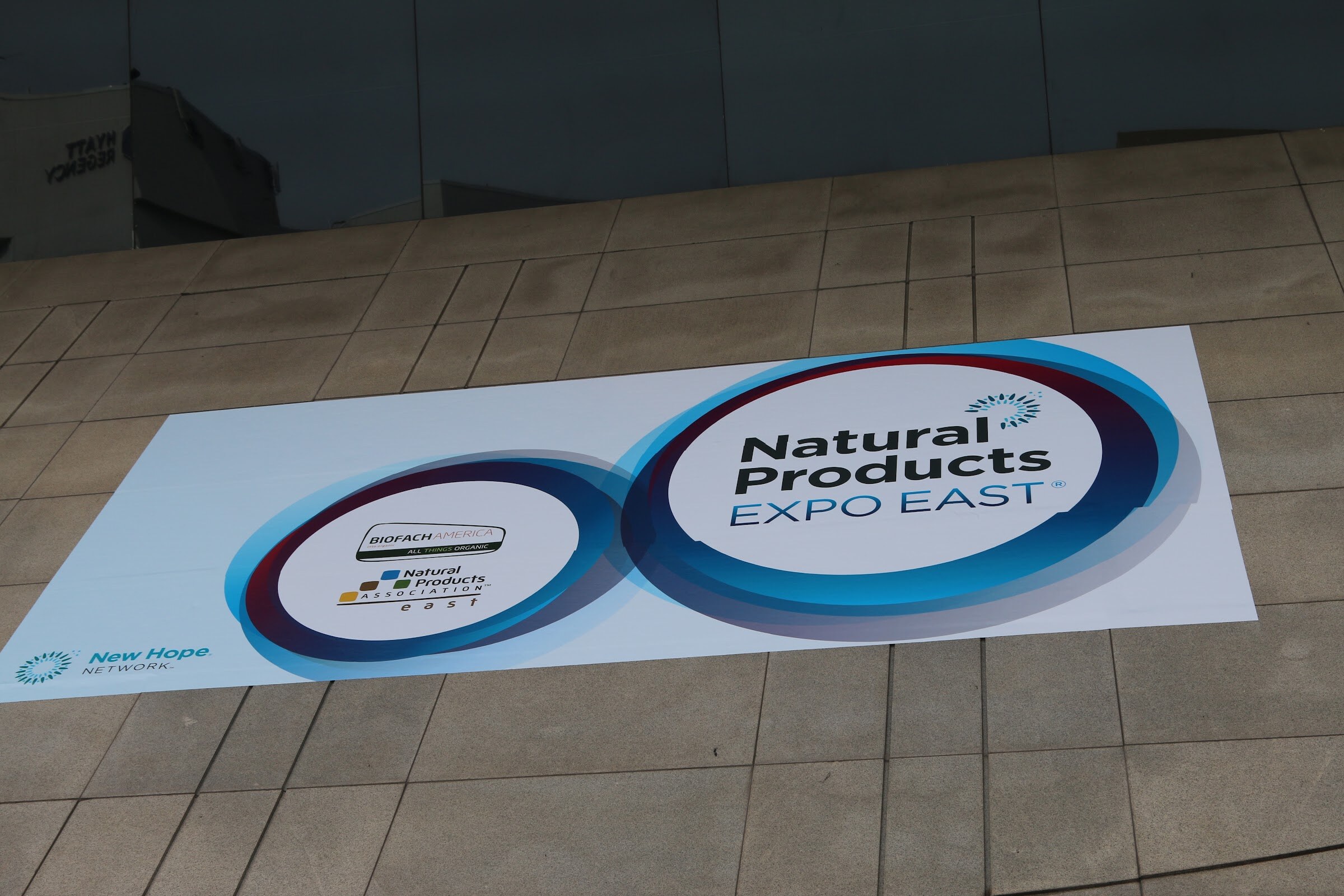 A white floor decal printed with the blue Natural Products Expo East logo and a small New Hope Network logo on gray tile.