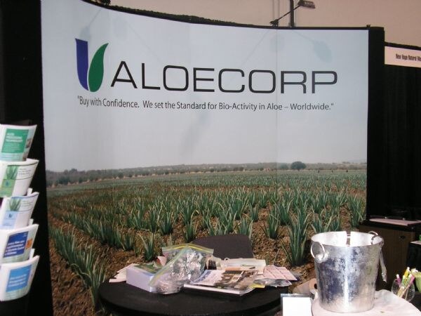 An Aloecorp booth with a curved backdrop showing a field of aloe plants and the tagline Buy with Confidence. Brochures sit in a rack at left and a table holds papers and an ice bucket.