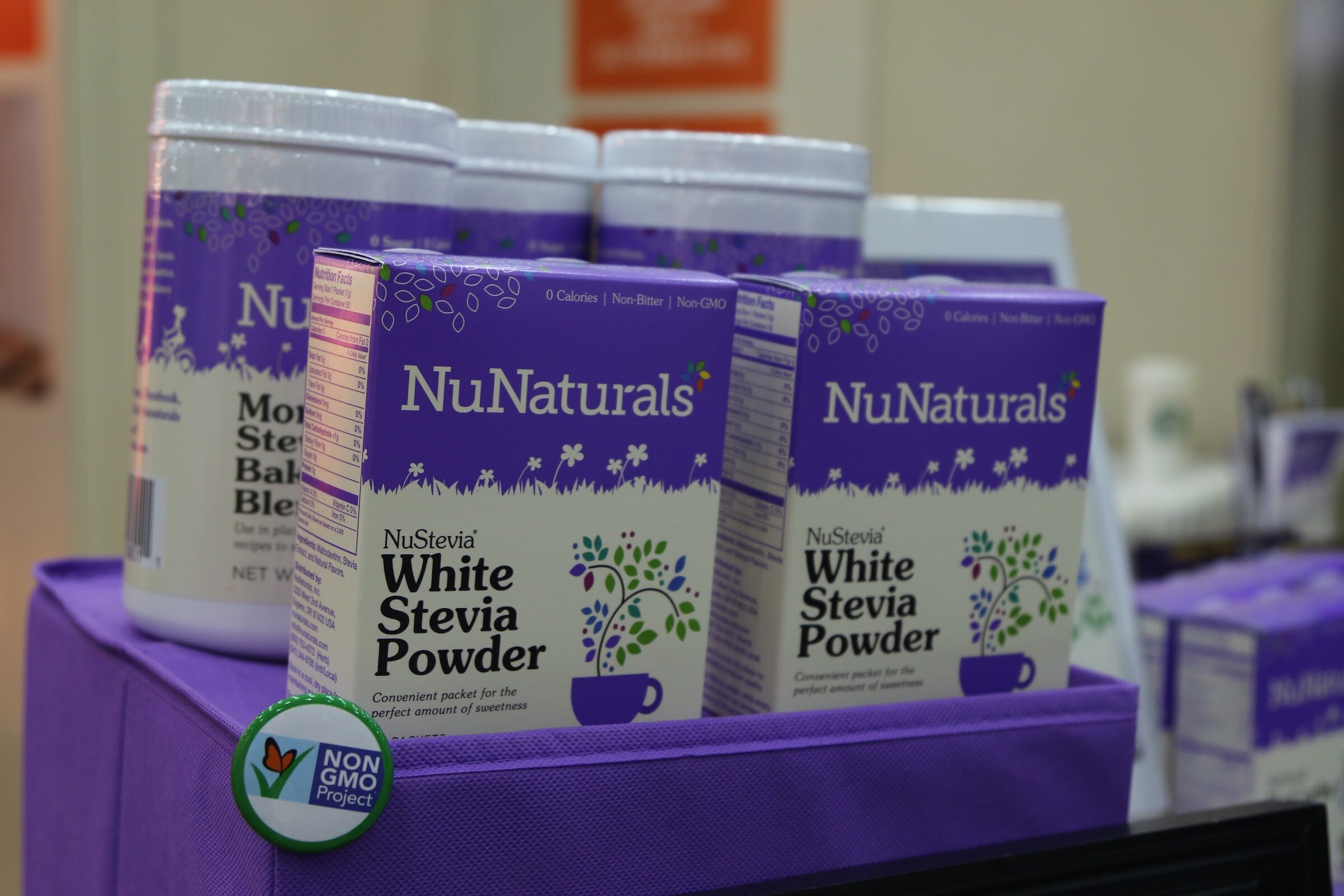 Two purple and white NuNaturals White Stevia Powder boxes in a purple fabric bin with a Non-GMO Project button at the front.