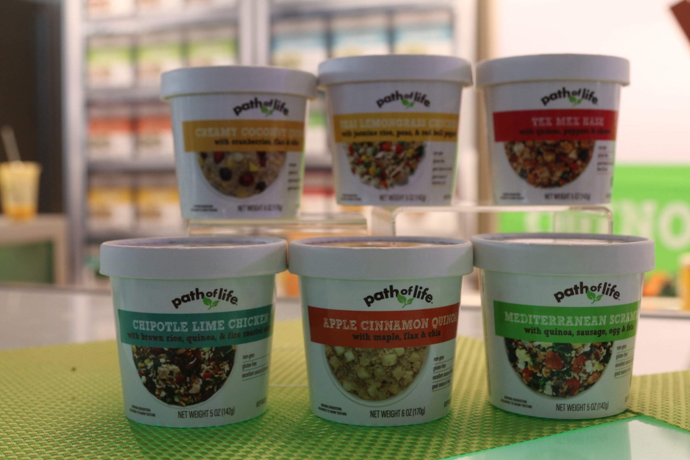 Six Path of Life cups with colored labels arranged in two tiers on a bright green mat with a refrigerated case behind.