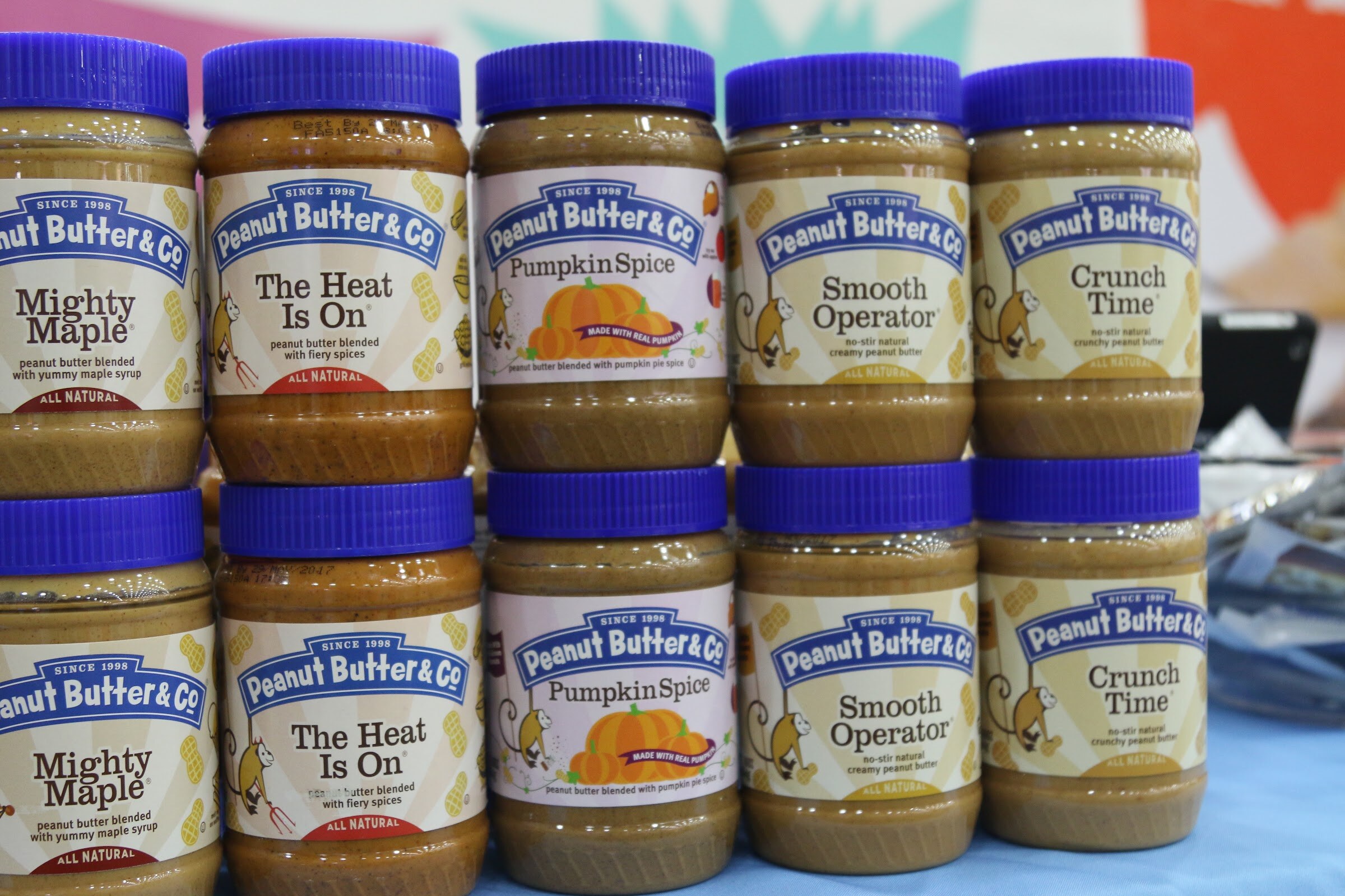 Two rows of Peanut Butter and Co jars with blue lids and monkey labels stacked on a blue tablecloth.