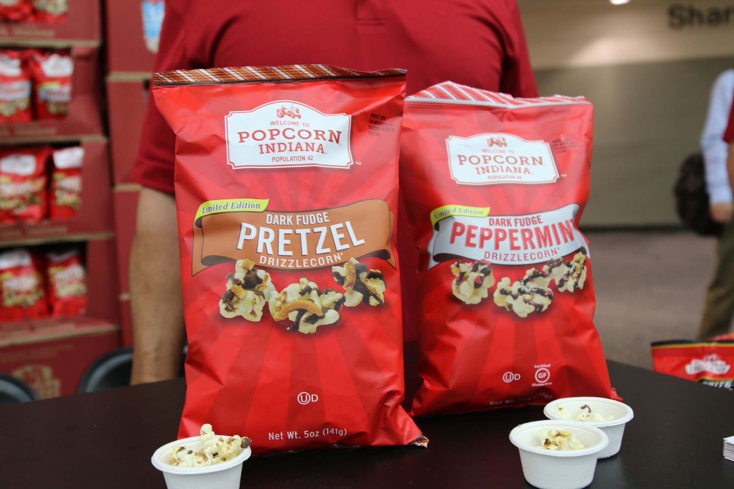Red Popcorn Indiana bags labeled Dark Fudge Pretzel and Dark Fudge Peppermint with two cups of popcorn samples on a table.