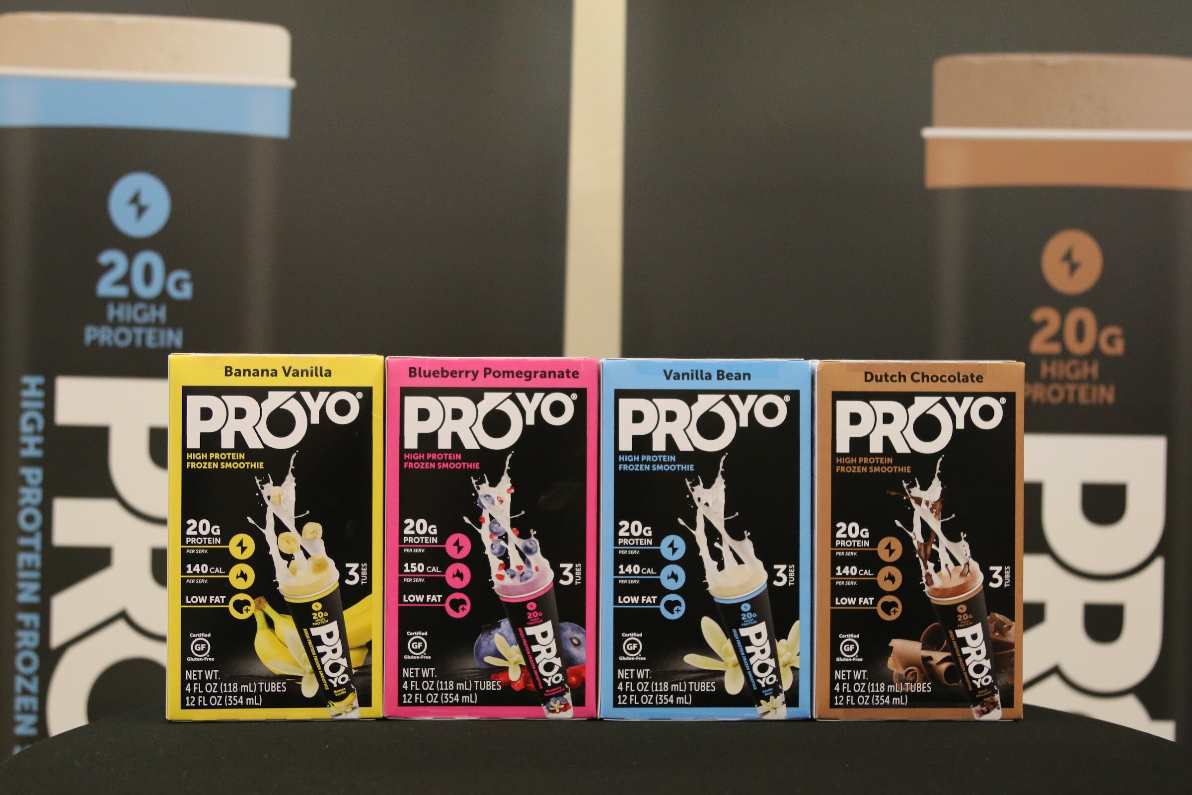 ProYo frozen smoothie boxes in banana vanilla, blueberry pomegranate, vanilla bean and dutch chocolate on a black table with a large 20g high protein banner behind.