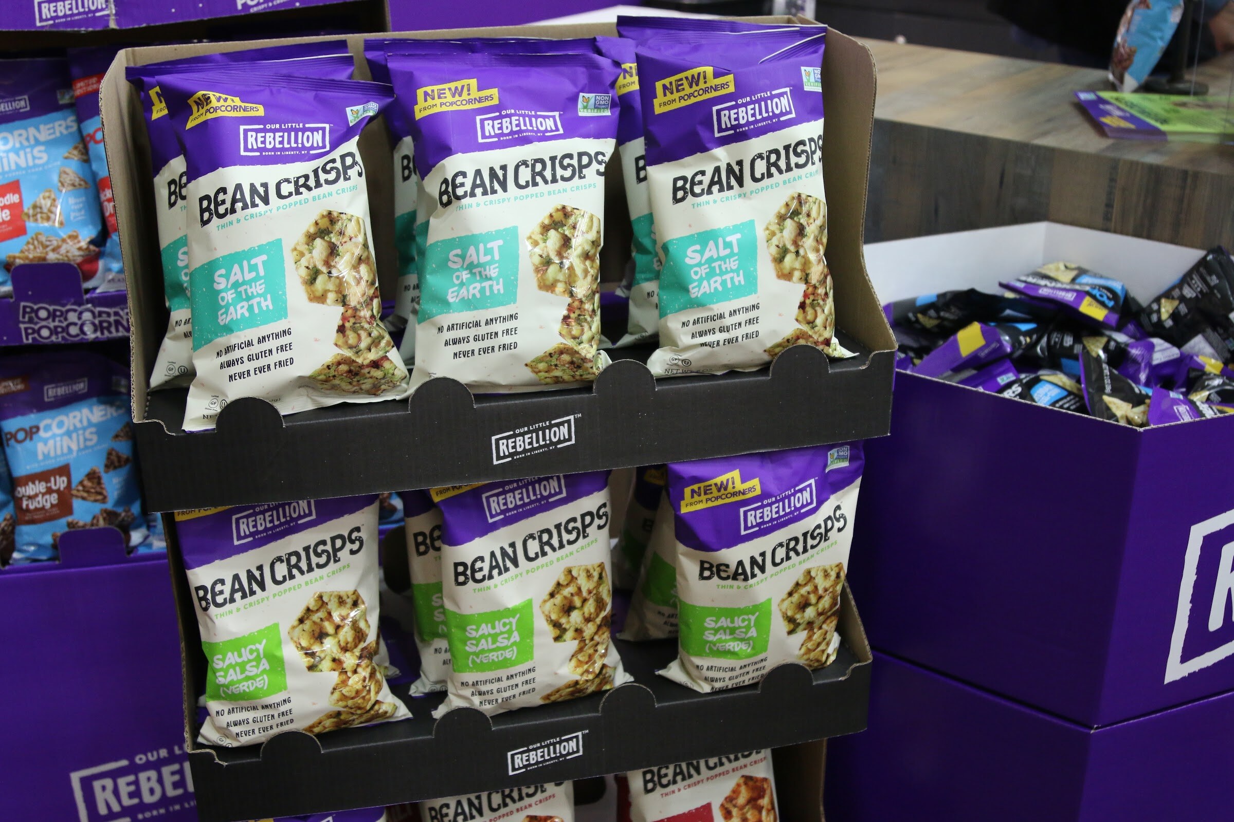 Purple Rebellion Bean Crisps bags stacked in a black cardboard display above a purple box.