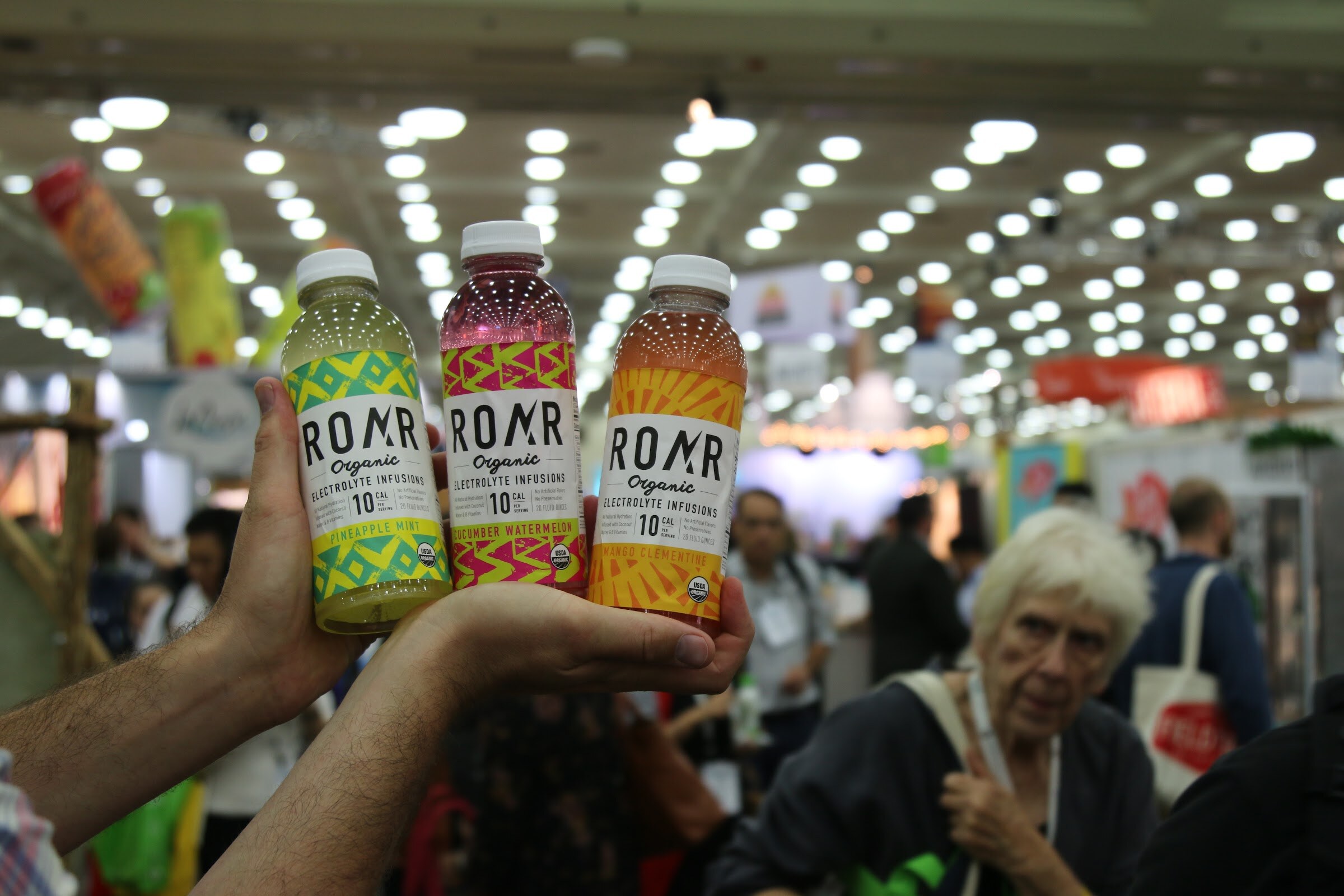A hand holding three ROAR Organic bottles in lime, berry and orange labels, with attendees and hanging lights out of focus behind.