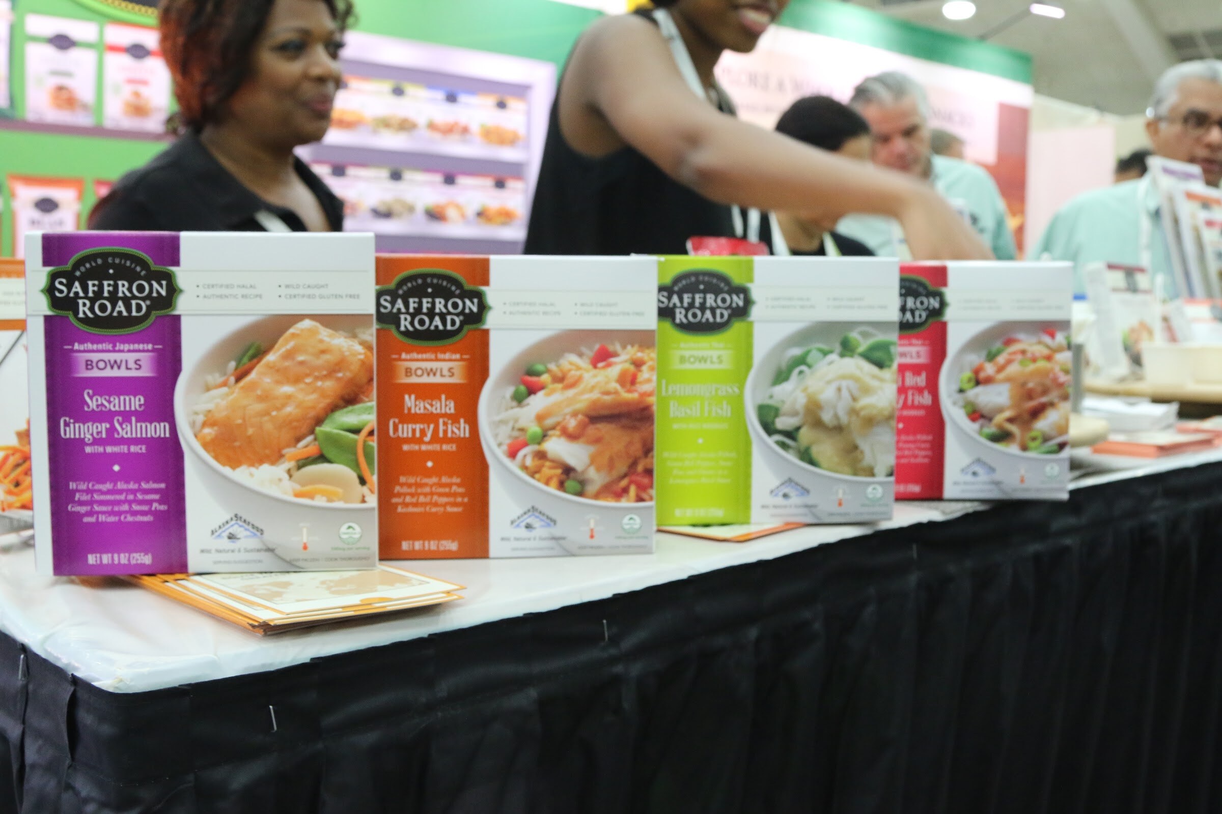 Saffron Road bowl boxes in sesame ginger salmon, masala curry fish and lemongrass basil fish on a table, with smiling booth staff blurred behind.