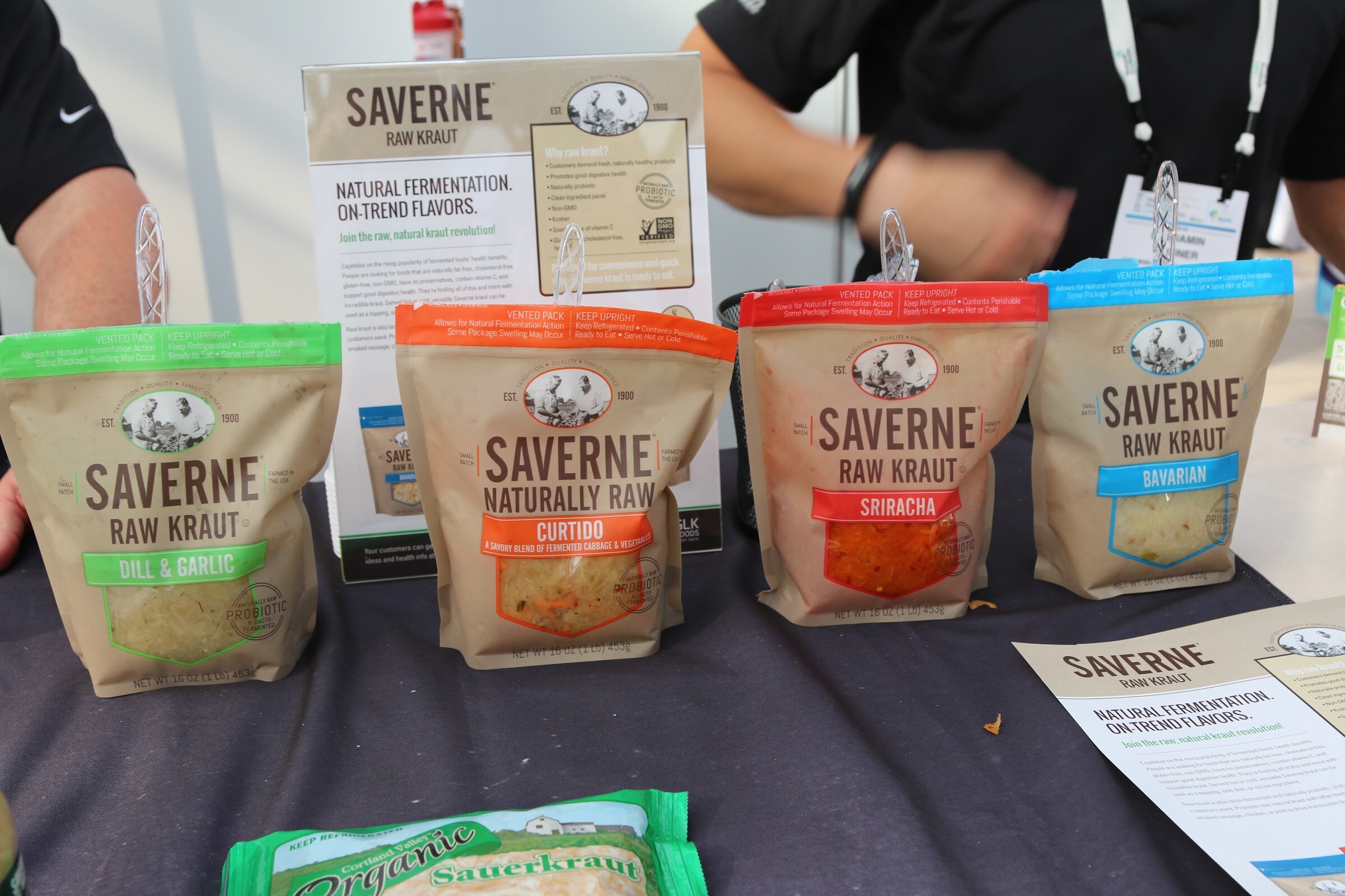 Four Saverne raw kraut pouches in dill and garlic, curtido, sriracha and Bavarian flavors on a dark table with an exhibitor behind them.