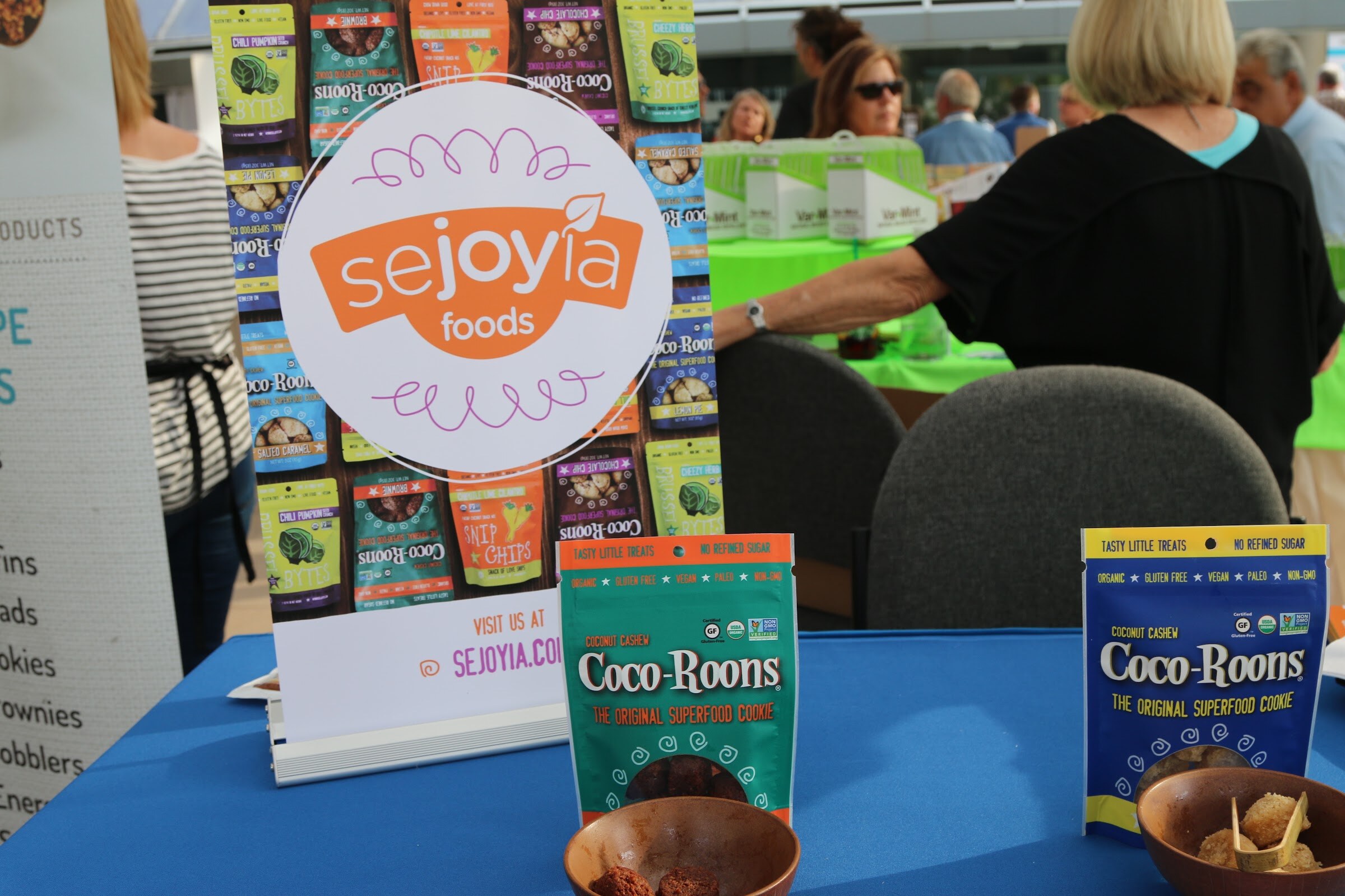 Two bags of Coco-Roons and small bowls of samples on a blue tablecloth under a round Sejoyia Foods logo, with attendees at tables behind.