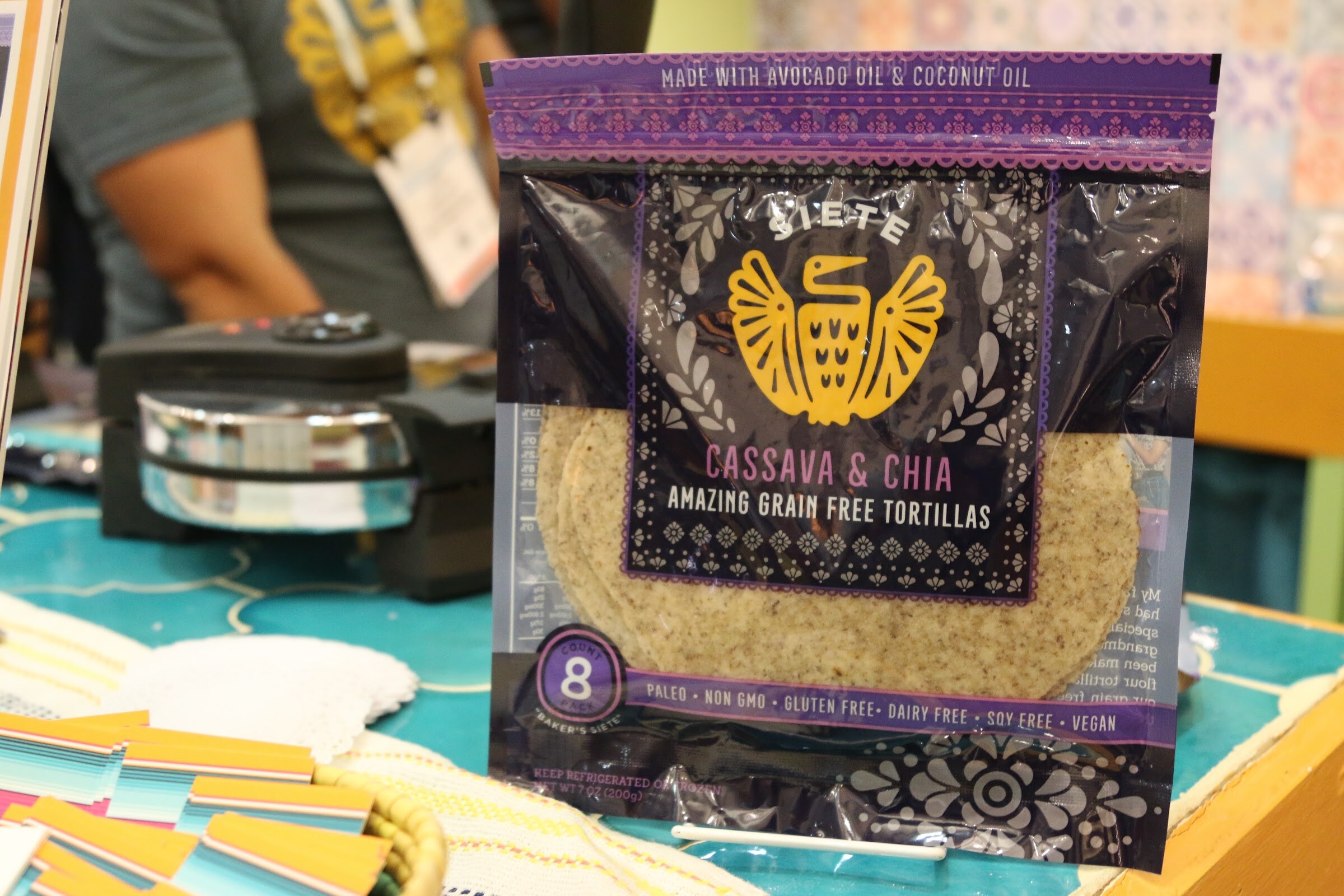 A purple Siete Cassava and Chia grain-free tortillas bag with a gold bird logo, a waffle-style press at left and an exhibitor in the background.