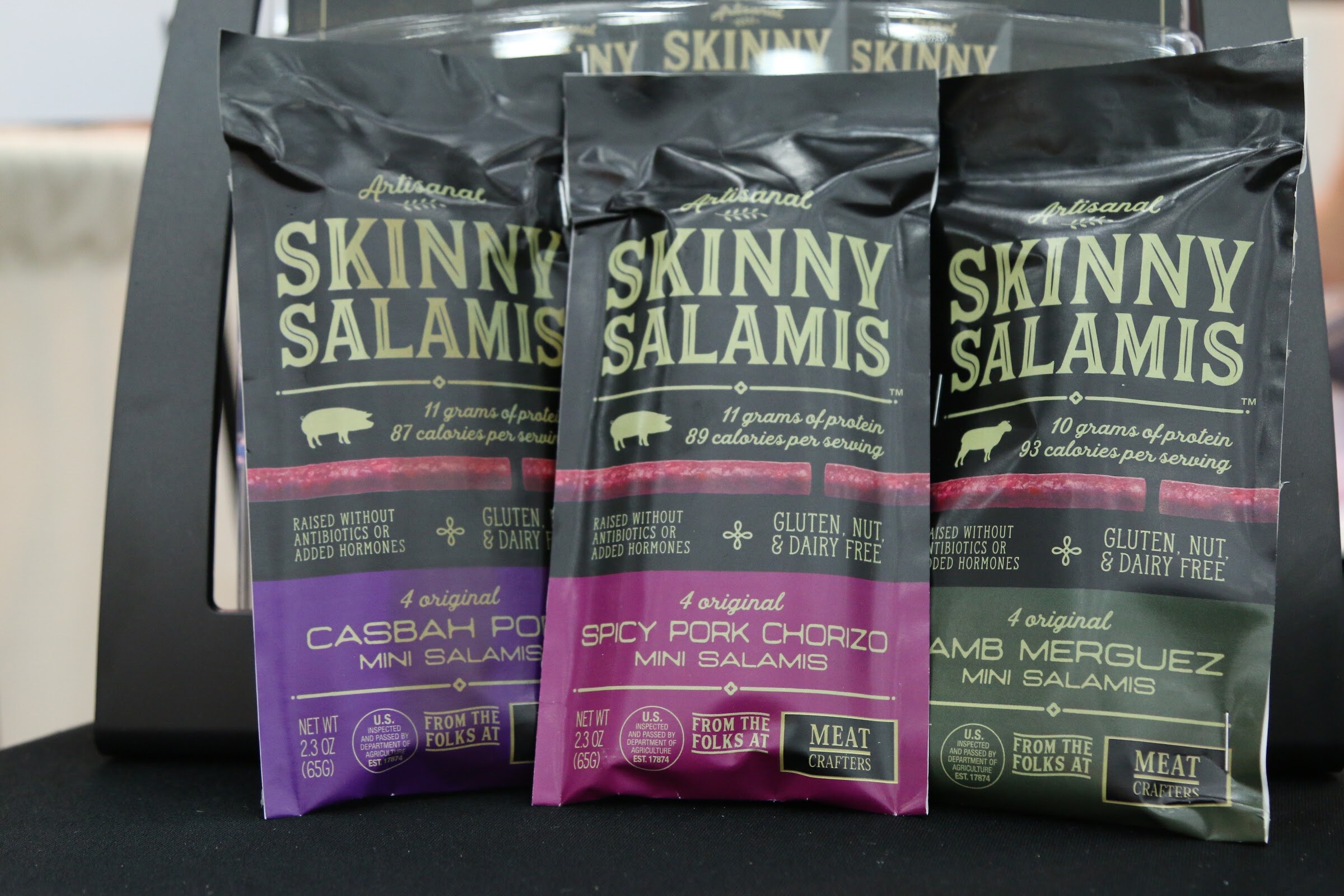 Three Skinny Salamis pouches in purple, pink and green flavors standing on a black table.