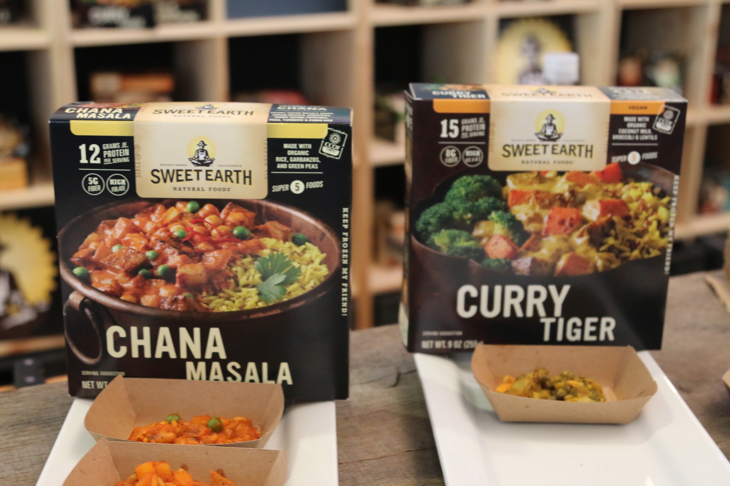 Two black Sweet Earth frozen meal boxes with food photos, with kraft paper trays of the dishes in front and wooden shelves behind.