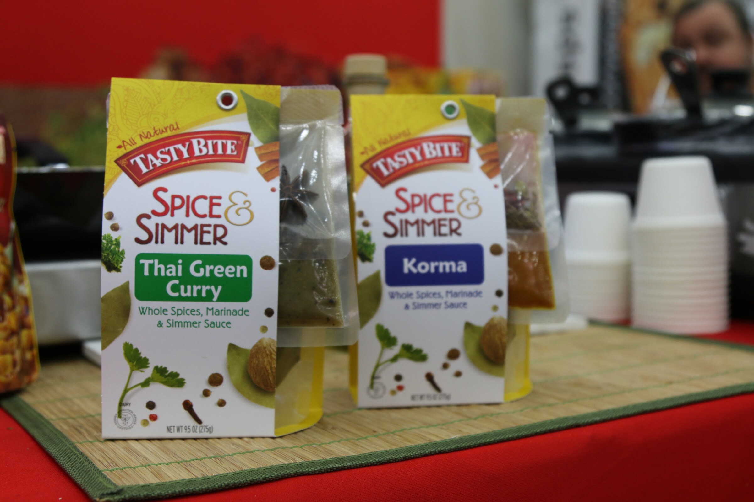 Two Tasty Bite Spice and Simmer packages in front of sample cups on a red tablecloth.