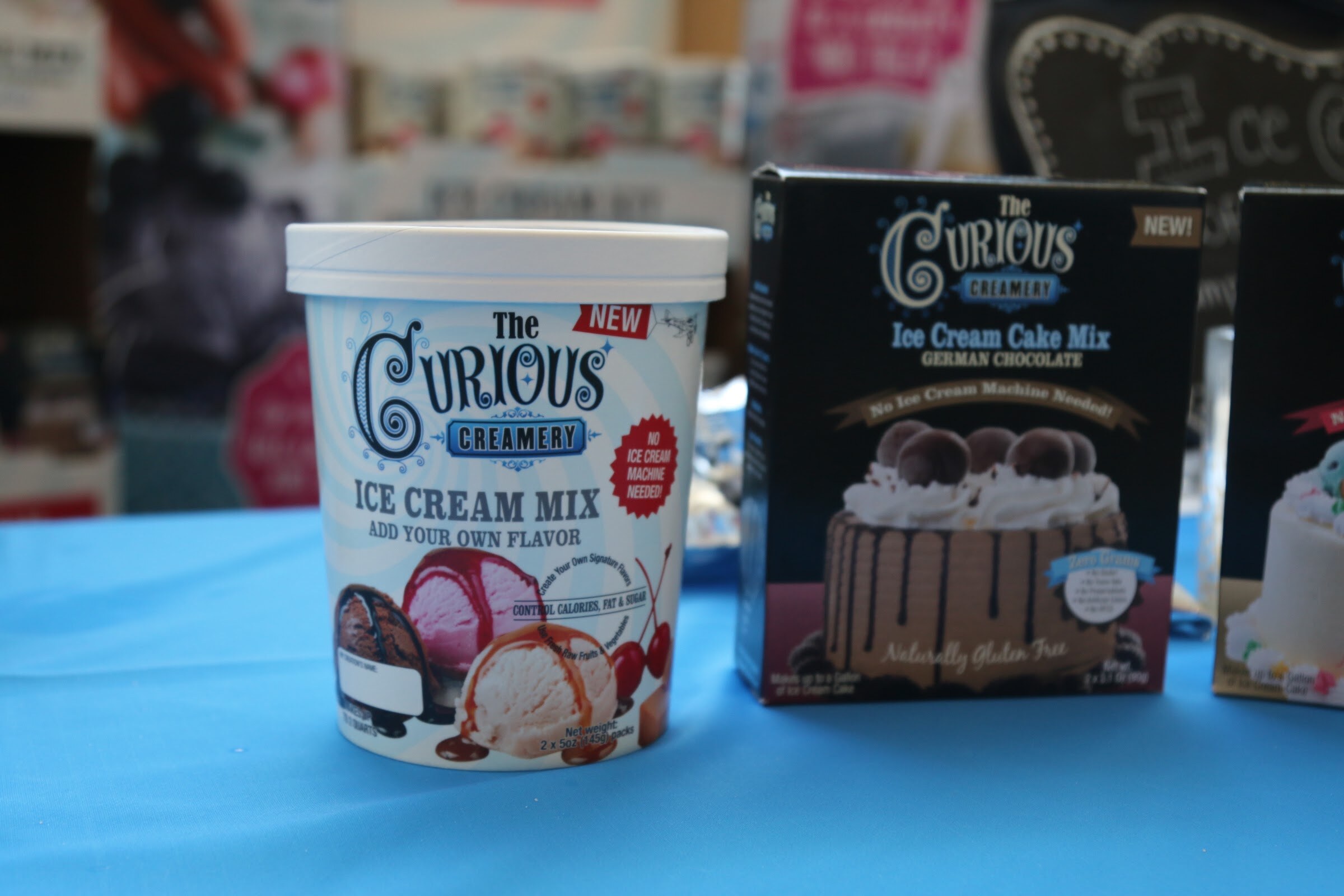 A white Curious Creamery tub with a cherry sundae photo next to a dark box of ice cream cake mix on a bright blue tablecloth.