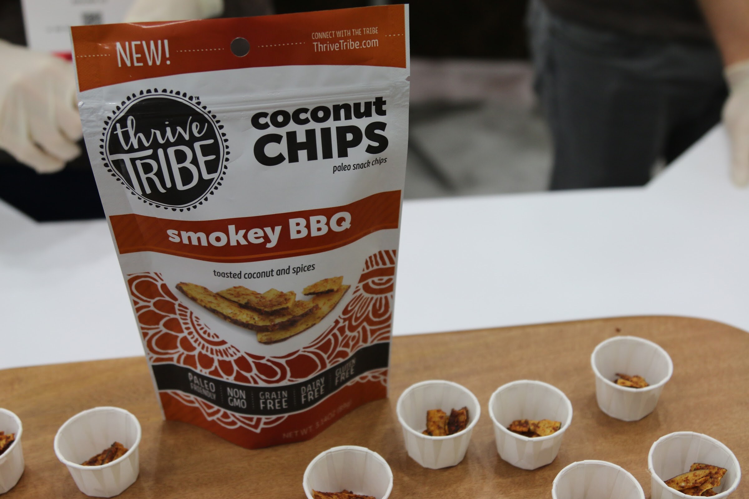 An orange Thrive Tribe coconut chips bag on a wooden board with several small paper cups of chip samples around it.
