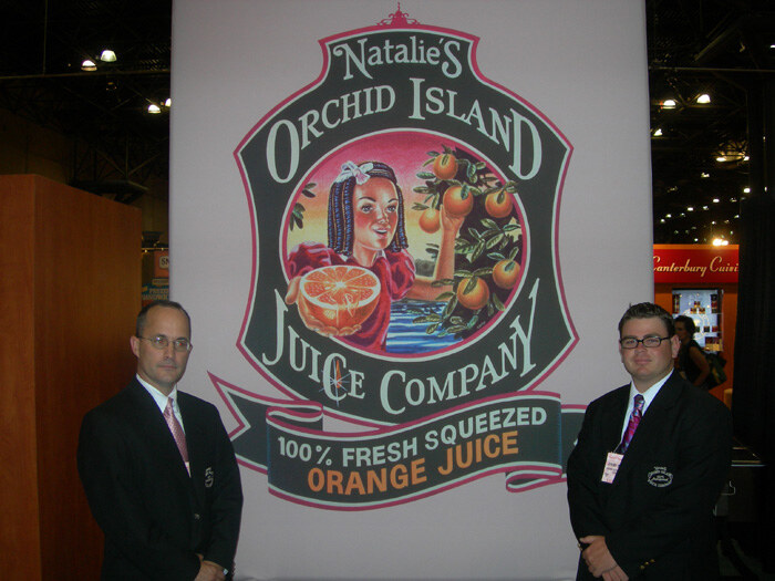 Two men in dark suits stand on either side of a large Natalie's Orchid Island Juice Company banner showing an illustrated girl holding an orange and reading 100% fresh squeezed orange juice.