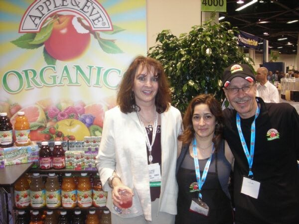 Two women and a man in a cap pose together in front of an Apple & Eve Organic sign and a shelf of Apple & Eve juice bottles and boxes. One woman holds a glass of pink drink.