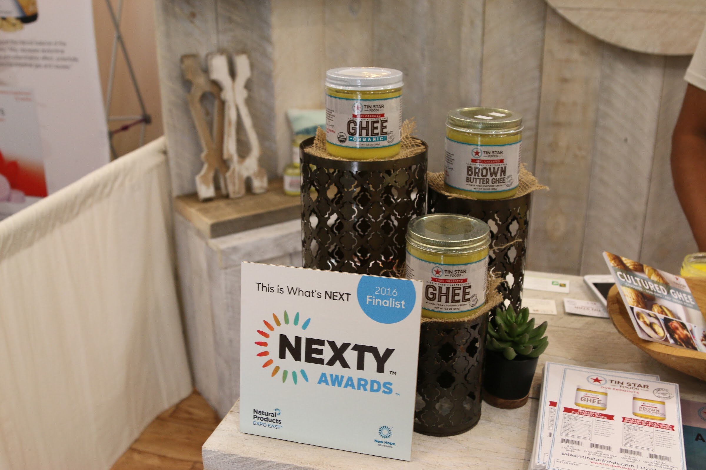 Three Tin Star ghee jars on dark metal stands in a booth with whitewashed wood walls and a NEXTY Awards 2016 finalist card.