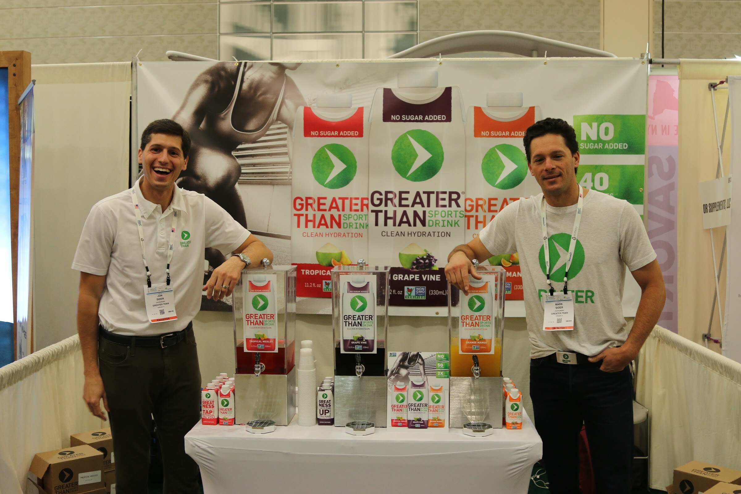 Two men with badges stand on either side of a table with drink dispensers in front of a Greater Than banner for clean hydration.